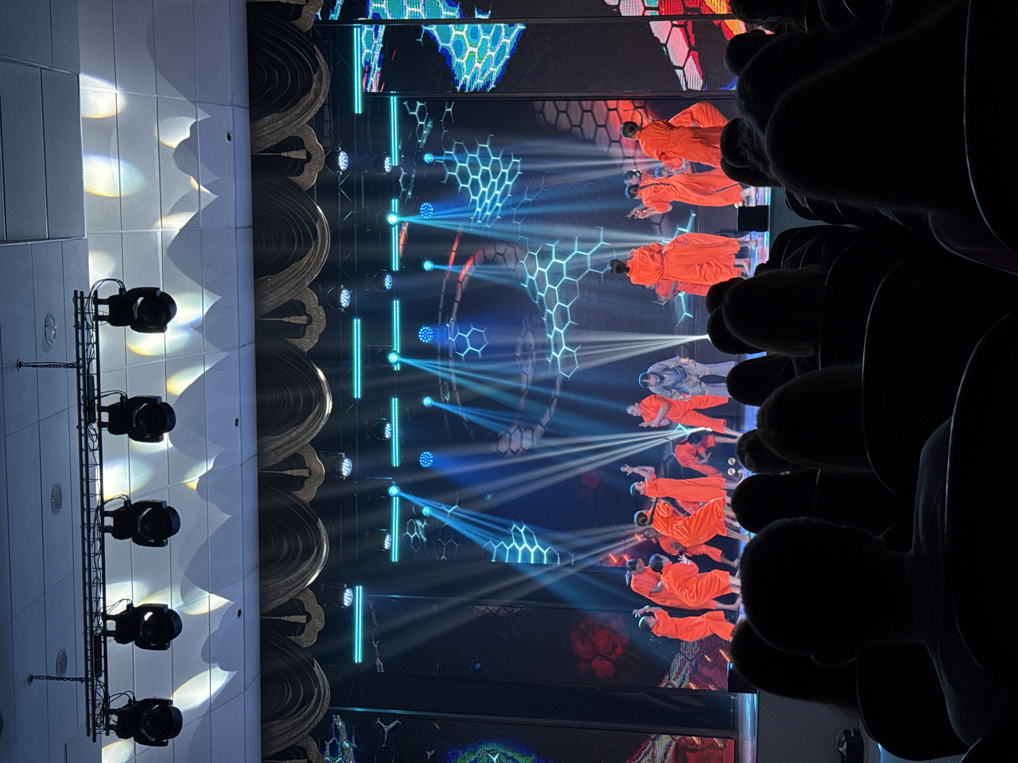 The image depicts a performance on a stage, likely within a theater or auditorium. Performers dressed in orange and silver attire are visible on stage with blue lighting and hexagon patterns displayed on the background screen. The view is from the audience perspective; silhouettes of audience members in the foreground. Stage lighting equipment is visible to the side, and ornamental architectural details are seen. Given the location in the city of Krasnoyarsk, Russia, the specific venue or event cannot be identified from the image itself.