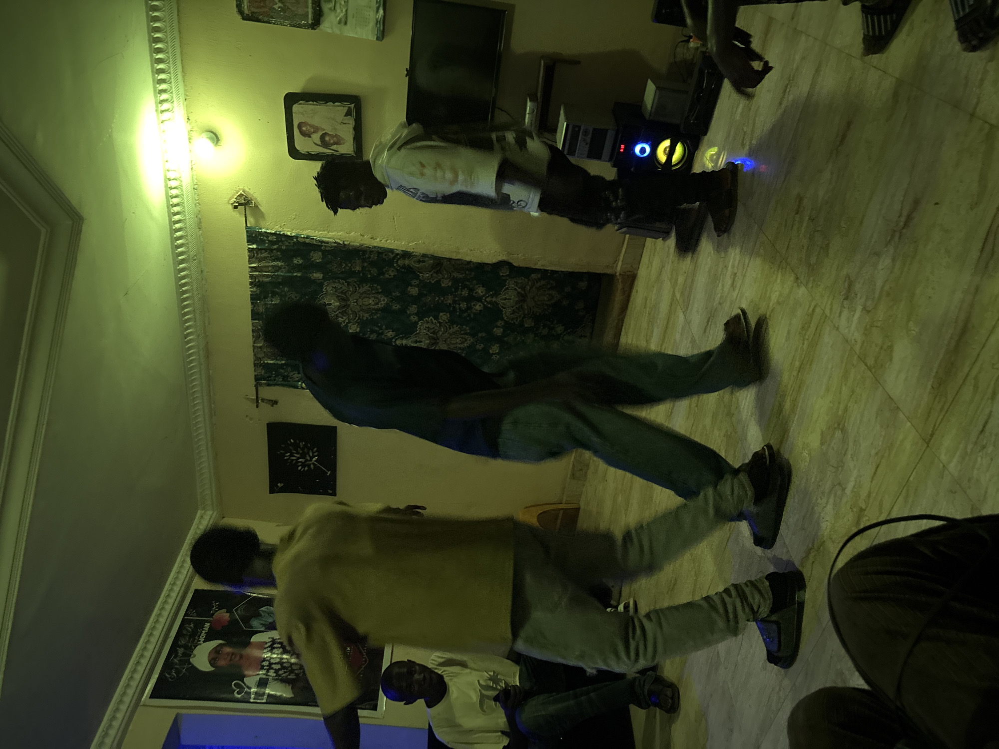 The image depicts an indoor gathering, possibly a party, in a room in Jos, Nigeria. The lighting is dim with a green hue, suggesting artificial lighting. Several individuals are present. In the foreground, the legs and lower bodies of two people are visible, one wearing dark pants and the other light-colored pants and dark shoes. To the left, a man in a light-colored t-shirt and jeans is seated, looking towards the center of the room. In the center, a person wearing a yellow shirt and light grey pants is captured mid-motion, possibly dancing or walking. To their right, another person in a dark t-shirt and jeans is also in motion, facing away from the camera. Further to the right, a person in a white hoodie with dark accents and dark pants is standing and looking to the side.
The environment appears to be a living room or communal space. On the wall to the left, a large poster featuring a woman in a headscarf is visible. Above it, decorative ceiling molding is present. To the right of the poster, a dark framed artwork is hung on the wall. Further to the right, green curtains with a floral pattern are drawn. Above the curtains, a light fixture illuminates the ceiling. On the far right wall, two framed pictures are displayed. Below the pictures, a television screen is mounted on the wall. Beneath the television, a shelf holds electronic equipment, including a stereo system with illuminated lights on the front. The floor is tiled with a marble-like pattern. The overall atmosphere seems relaxed and informal, with people engaged in social interaction.