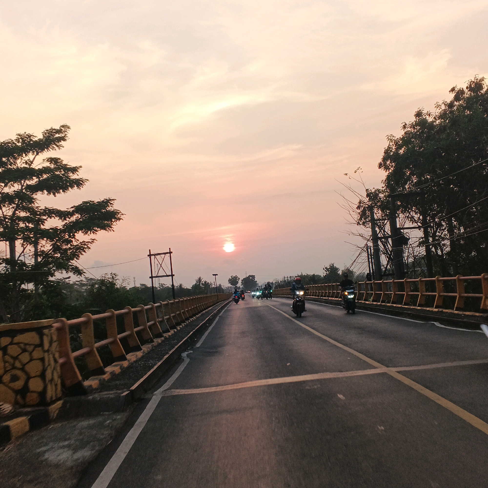 A view of a bridge in Kalikudi, Indonesia, during sunset. The sky is filled with soft orange and pink hues, with the sun partially obscured by clouds. A road stretches across the bridge, with several motorcycles riding in the same direction. The bridge has a yellow railing with stone-like structures on the left side. Trees line the sides of the road and the bridge, with some power lines visible in the distance. The overall scene depicts a tranquil evening commute.