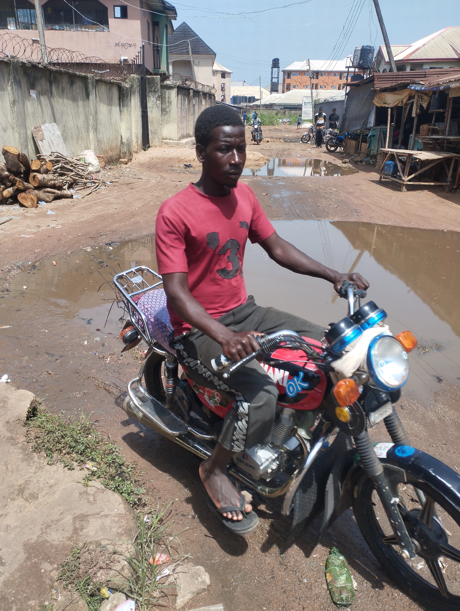 A man on a red motorcycle is pictured in a flooded dirt road in Eziobodo, Nigeria. He wears a red t-shirt, dark pants, and sandals. Behind him is a pile of wood on the left, buildings with barbed wire, and a partially visible street with a water tower, other buildings, and a market stall to the right. Other motorcycles are seen in the distance. Debris is scattered on the ground.