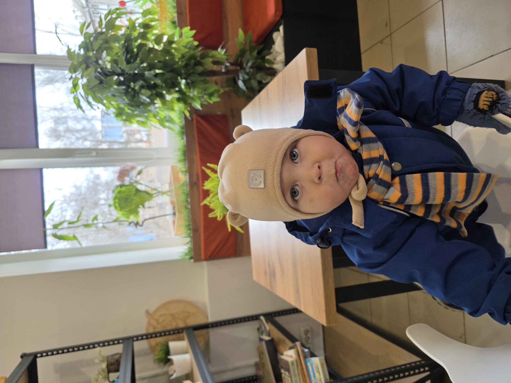 A baby with blue eyes is sitting at a wooden table in what appears to be a cafe or restaurant. The child is wearing a beige hat with ears, a blue jacket, and a striped scarf. The baby is holding something in their gloved left hand.
Behind the baby is a window with plants, and a black metal shelving unit with various objects on it, including books and plants. The scene suggests an indoor setting, possibly in the city of Chaikovsky, Russia, given that was specified.
