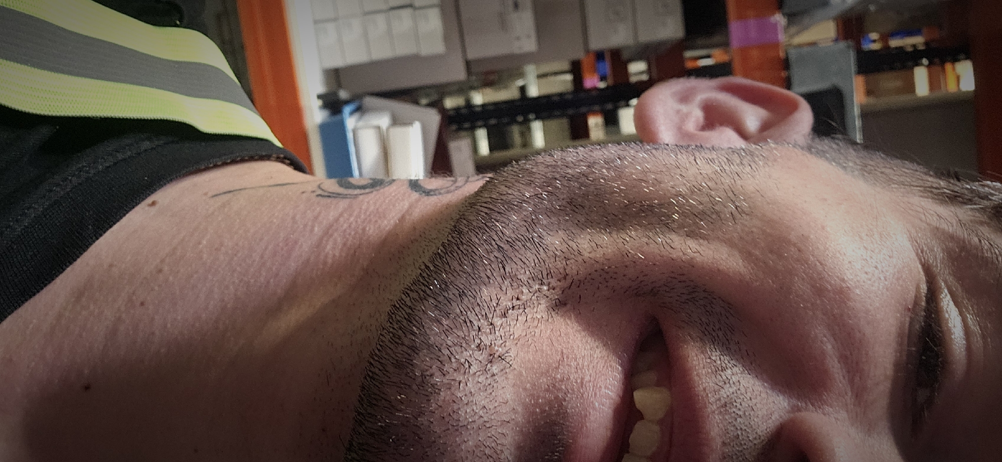 The image presents a close-up, angled view of a person, likely male, appearing to be resting or lying down. The shot focuses on the individual's face, specifically the forehead, cheek, and chin, with one eye partially closed and a slightly open mouth revealing teeth. The person has short, dark stubble on their face and hairline, with individual hairs showing glints from the lighting. A prominent ear is visible in the upper right quadrant of the image.
On the person's neck and shoulder, a tattoo with abstract, circular patterns in black ink is visible. They are wearing a dark garment, possibly a t-shirt, underneath a bright yellow/lime green and grey striped strap or band, suggesting it could be part of a safety vest or work attire.
The background is out of focus but indicates an indoor, possibly industrial or storage environment. Blurred shapes suggest shelving units or racks holding various items like boxes and containers, some appearing white or blue. The lighting seems artificial. No specific emotions are clearly conveyed, but the posture suggests relaxation or a moment of rest. There is no visible text in the image. While the image itself does not provide explicit location cues, the provided context indicates the location is Plovdiv, Bulgaria.