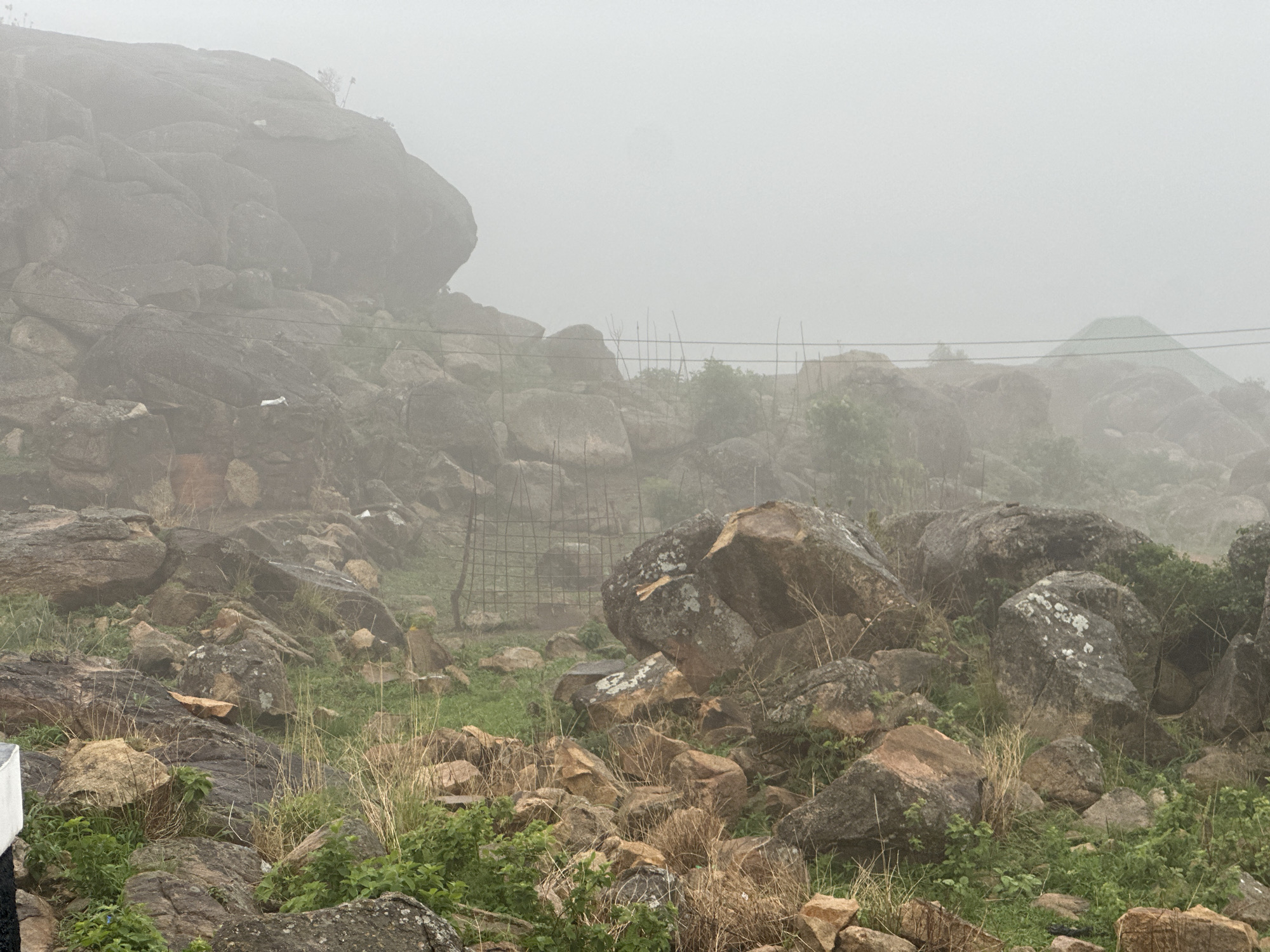 The image shows a rocky, hilly landscape obscured by heavy fog. The foreground is filled with various sized rocks, patches of green vegetation, and dry grasses. A crudely constructed fence-like structure made of sticks and wire netting is visible in the middle ground. A large, imposing rock formation dominates the upper left, almost entirely shrouded in fog. Power lines stretch across the frame in the upper portion of the image. The overall scene appears to be in a rural or mountainous area. It is confirmed that the location is Jos, Nigeria.
