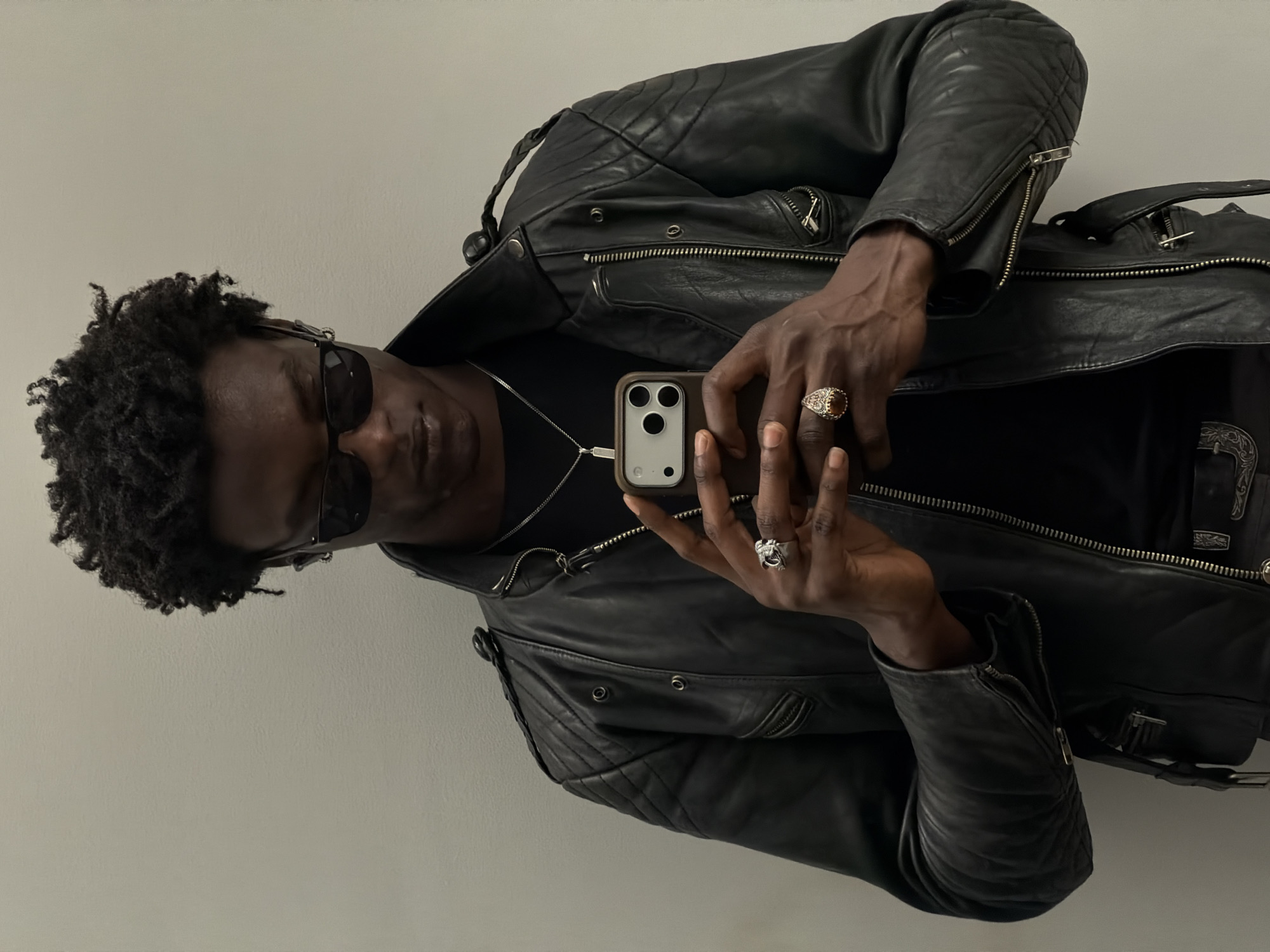 A dark-skinned man with short, curly hair and sunglasses is holding a phone with both hands. He is wearing a black leather jacket over a black shirt and a silver chain with a pendant. He also has two rings on his fingers. The background is a plain, light-colored wall. The location appears to be a studio or indoor setting, not identifiable as London.