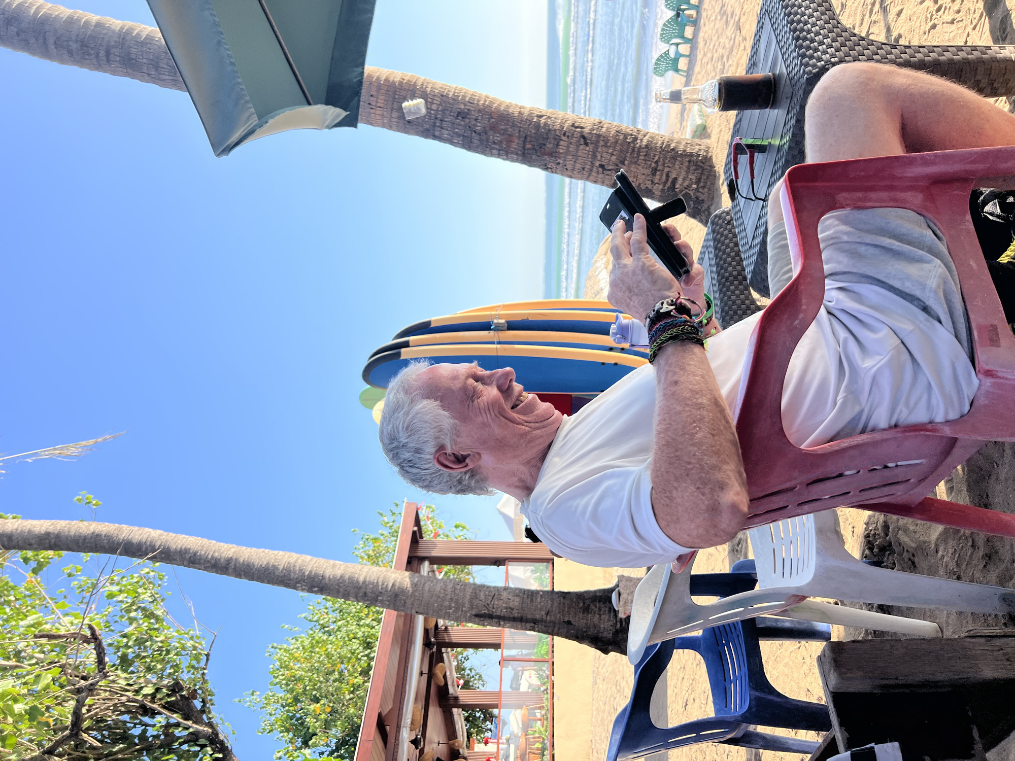 An elderly Caucasian man with gray hair is sitting in a red plastic chair on a sandy beach. He is wearing a white t-shirt and light-colored shorts. He is holding a black smartphone in his hands and looking at it, smiling. He has several bracelets on his left wrist. A black table is in front of him with a bottle of dark liquid and a pair of red-framed glasses on it. Behind him, there are several surfboards leaning against each other, a palm tree, and the ocean with waves. In the background, there is a beachside establishment with a roof and tables, and more palm trees. The sky is clear and blue. The location appears to be Kuta, Indonesia, given the beach setting, surfboards, and tropical vegetation.