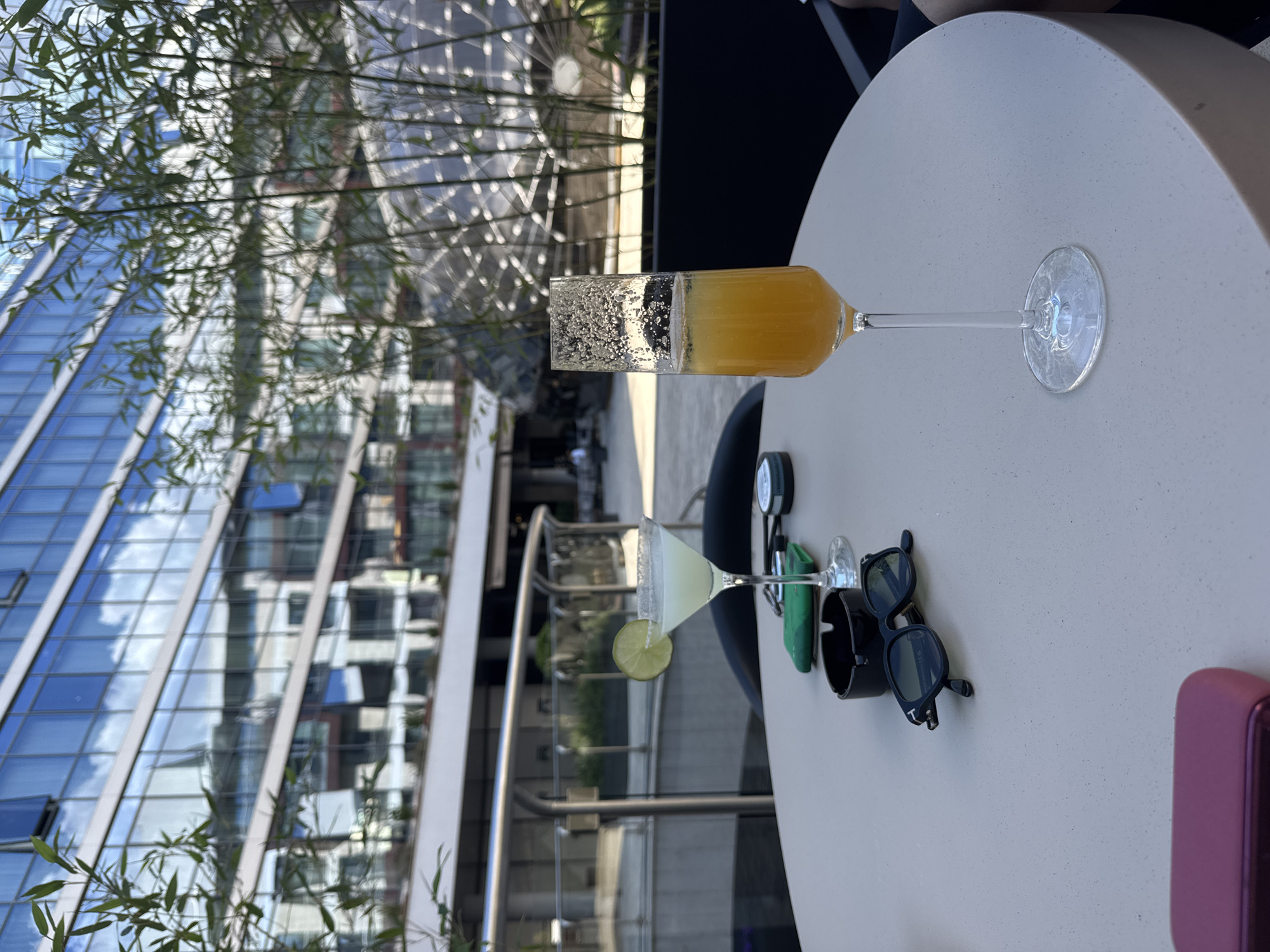 The scene captures an outdoor setting on a high-rise patio in Podgorica, Montenegro. The view includes a modern building with a blue glass facade and foliage in the foreground. Two cocktails sit on a round table. One is a margarita with a lime wedge, the other a layered cocktail with a foam topping and a yellow-orange liquid below. Nearby are sunglasses, an ashtray, and a pink object that is out of focus. The patio overlooks a building with architectural lines visible through the foliage. The atmosphere appears relaxed and upscale.