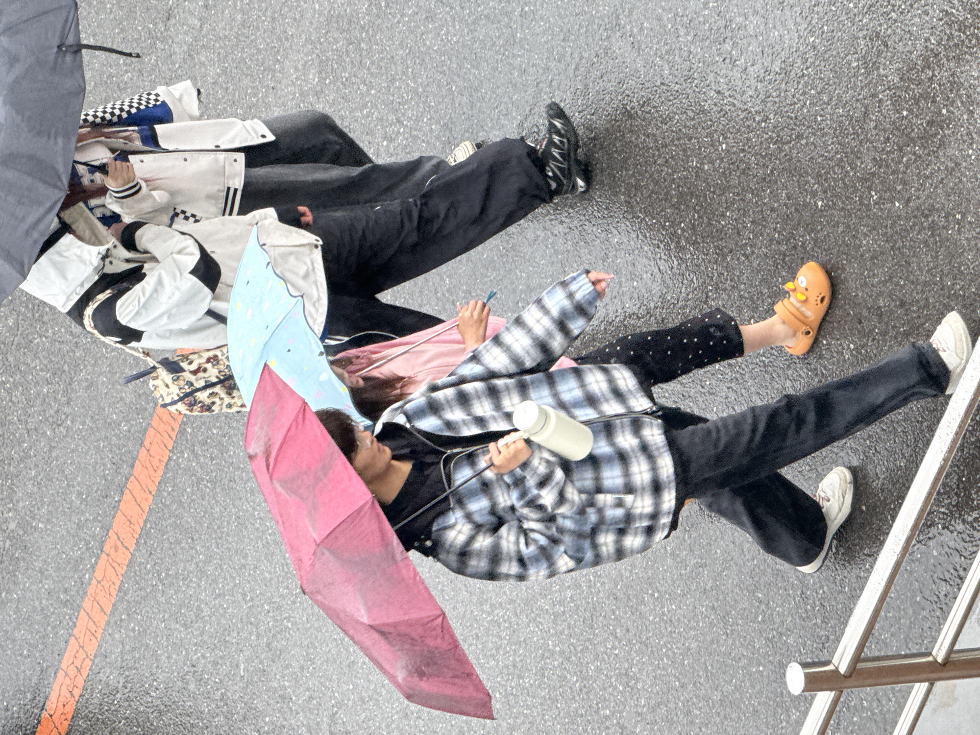 Aerial view showing people lying on a wet surface in Zhongtun, China, apparently during or just after a rain shower. The surface appears to be asphalt or concrete, with a reflective sheen. Two individuals are seen from above, with sections of a third person in view. The individual closest to the camera is holding a multi-colored umbrella with pink, blue, and white panels. They are wearing a plaid jacket, black pants, and white sneakers. Their left foot is adorned with an orange Croc-style shoe, decorated with yellow adornments. They are holding a cylindrical white object, perhaps a water bottle or similar item. Further away is another individual covered by a white jacket with black accents and black pants. Part of a third person is also visible under a black umbrella. An orange line is painted on the surface at the lower left of the frame, and metal railings appear at the lower right.