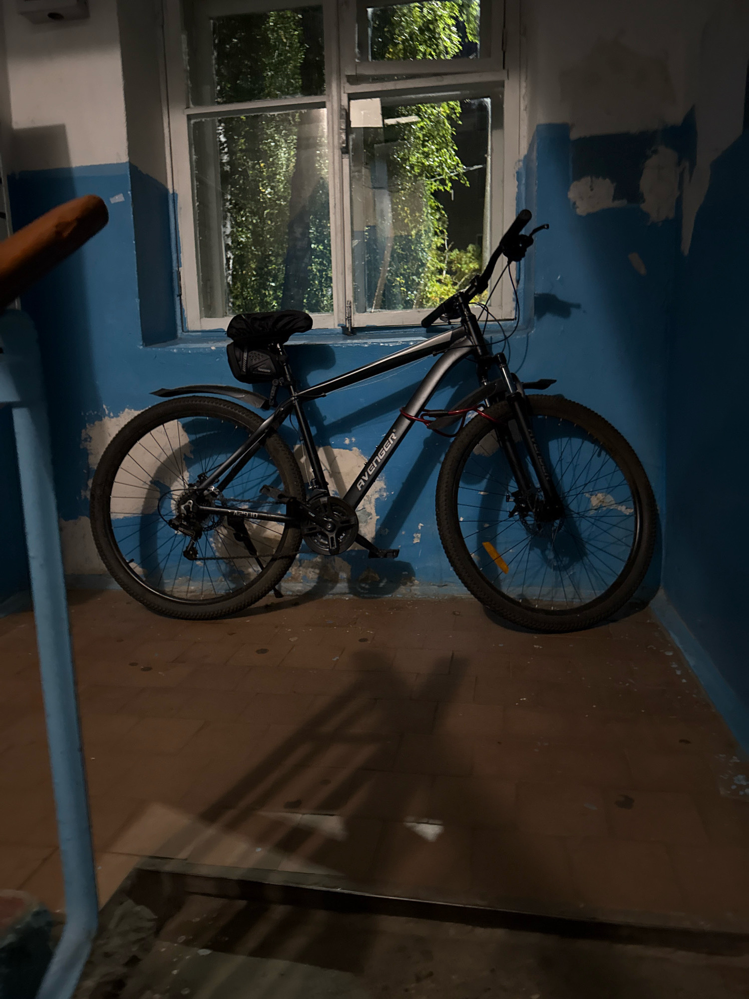 A black bicycle is parked indoors, leaning against a blue wall with peeling paint. A window with greenery visible outside is behind the bicycle. A blue handrail is in the foreground. The floor is tiled with brown square tiles. The lighting suggests it is evening or night. The scene appears to be in an apartment building hallway or staircase in Zavolzhye, Russia.
