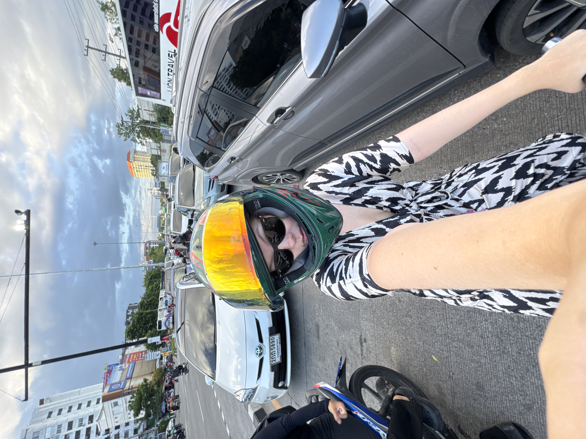 A woman wearing a helmet and sunglasses is taking a selfie while sitting on a motorcycle in traffic in Chiang Mai City Municipality, Thailand. She is wearing a black and white patterned dress. The traffic consists of cars and other motorcycles. In the background, there are tall buildings, trees, and a cloudy sky. A bus with "LION TRAVEL" written on it is visible. The motorcycle license plate has Thai characters. The ground is asphalt.