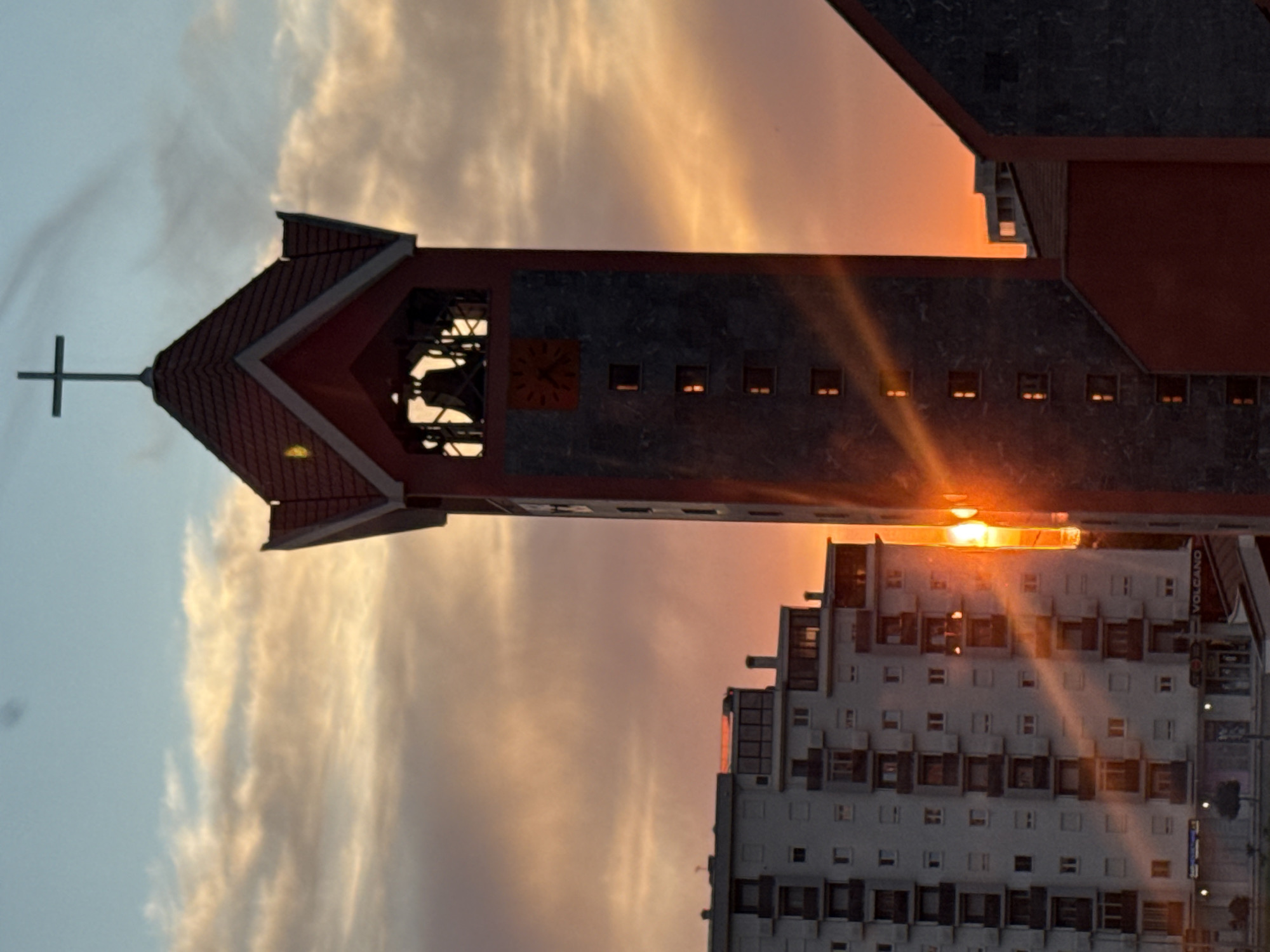 A vertical, slightly angled photograph captures a sunset scene featuring a church tower and an apartment building. The left side of the image reveals a sky with white and orange clouds. The church tower, positioned in the center, has a cross atop its peaked roof, an open bell tower, and a clock face. The tower's brickwork appears to be a combination of grey and red. The apartment building, on the right side of the frame, is white with symmetrical windows and dark window frames. Sunlight appears to be shining directly between the church and the building. A sign reading "VOLCANO" is visible on the side of the apartment building. Based on the architectural style and signage, the location is likely in Bar, Montenegro.
