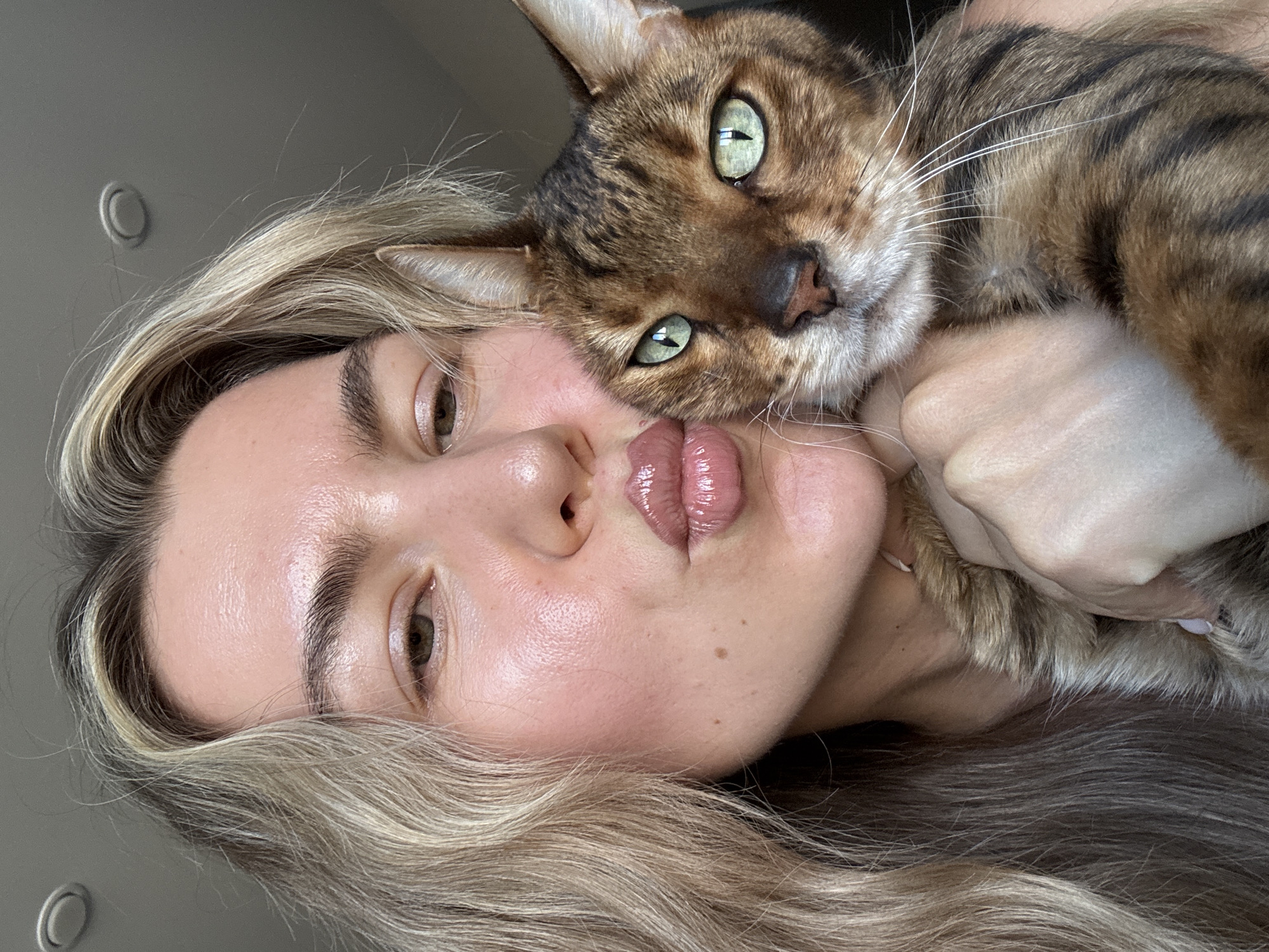 This close-up portrait features a woman and a cat in an intimate, vertical composition. The woman, positioned on the left, has long, wavy blonde hair with visible darker roots, and fair skin with a natural, glowing complexion. She looks towards the camera with her head tilted, making a playful "kissing face" with glossy, plump pink lips. Her eyes are light brown or hazel, and she has full, well-groomed eyebrows.
A Bengal cat is nestled close to the woman's face, resting its head on or near her cheek and shoulder. The cat is looking directly at the camera with piercing bright green eyes and prominent black pupils. Its fur is a distinctive pattern of brown, black, and tan stripes and spots, and long white whiskers are visible around its muzzle. One of the woman's hands is visible on the right, gently holding the cat.
The background is soft and out-of-focus, appearing as a plain, light grey wall. A subtle circular imprint or fixture is dimly visible on the wall in the upper left corner. The natural lighting suggests a daytime setting. The overall mood is affectionate and heartwarming, showcasing a strong bond between the woman and her pet. There is no visible text in the image.