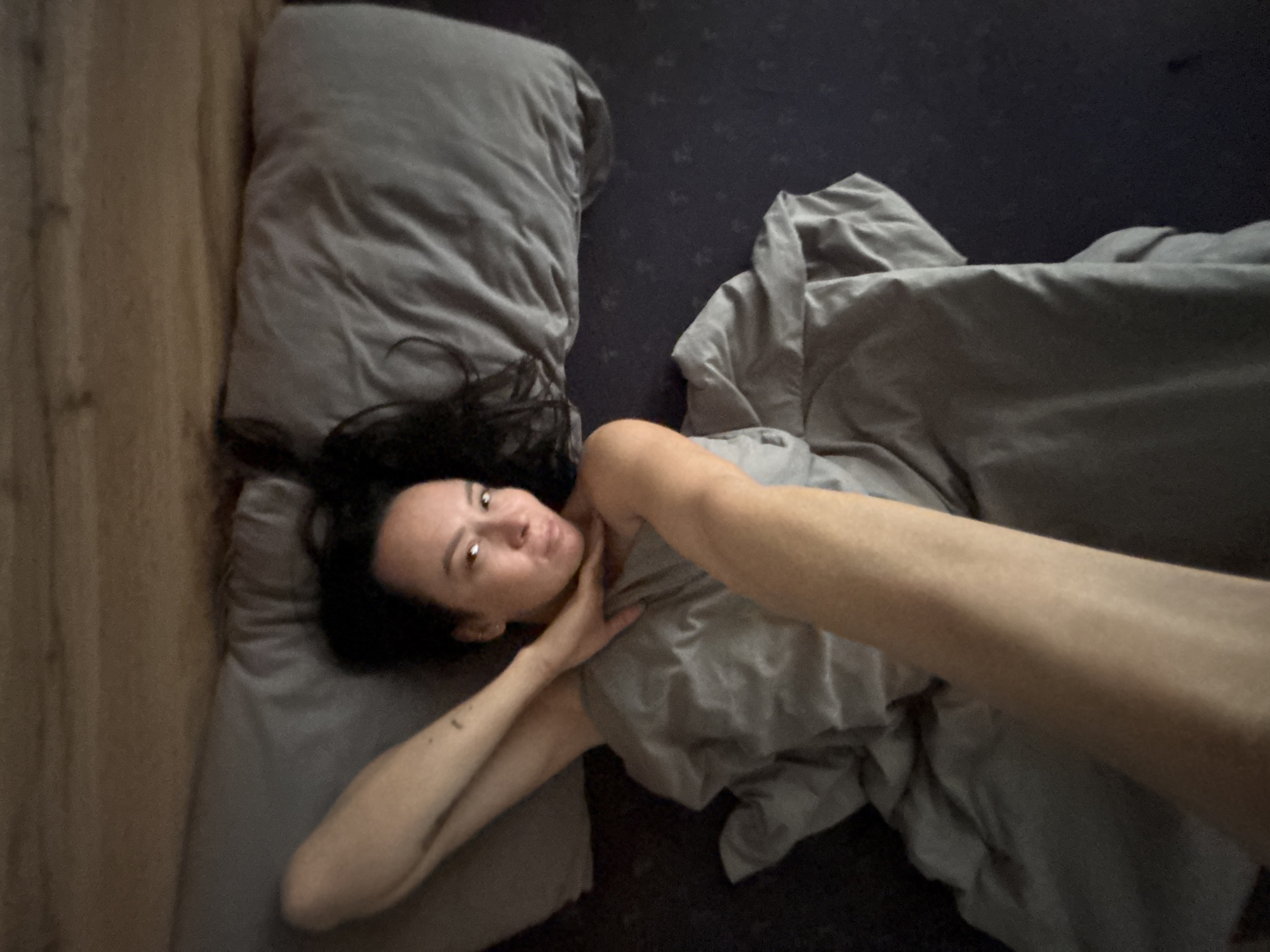 A woman lies in a bed with gray bedding. She is fair-skinned with dark hair. She is touching her neck with her right hand. The bed is positioned next to a wall with a light brown vertical surface, possibly wood paneling. The location in Chaikovsky, Russia is not discernable from the image.