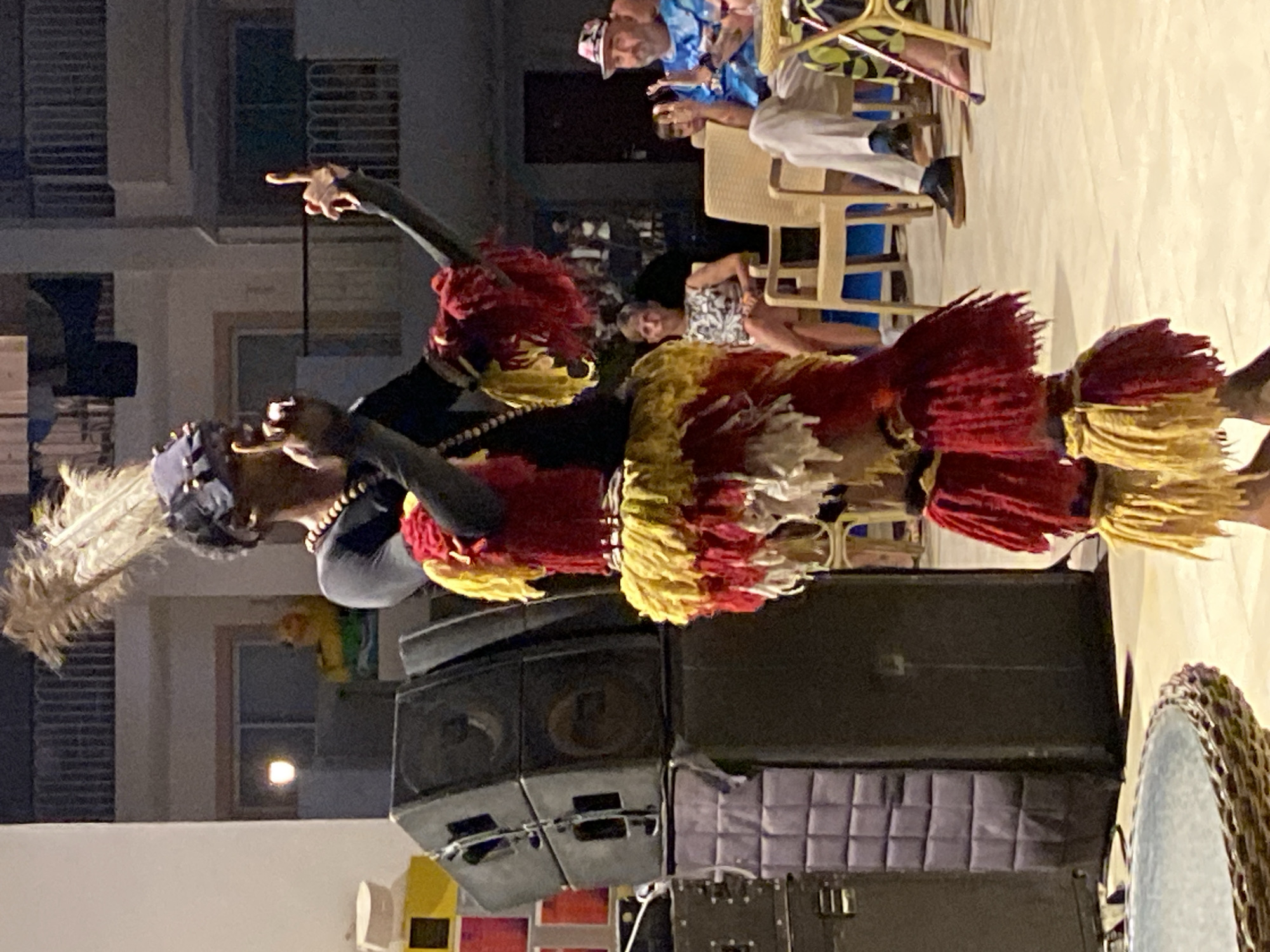 An outdoor evening scene in Sharm El Sheikh, Egypt, features a performer in traditional costume dancing in front of a stack of large black speakers and other electronic equipment. The performer wears a black long-sleeved shirt, a beaded necklace, a dark headpiece adorned with a large feather plume, and a vibrant, layered skirt of red, yellow, and white fringes. The performer's arms are raised, and their fingers are extended in a gesture. In the background, several people are seated on chairs at tables, appearing to observe the performance. One man is wearing a blue shirt with a colorful pattern and a patterned hat, and he is clapping. A woman to his left is wearing a black and white patterned sleeveless dress. The setting appears to be a resort or entertainment area, with a building structure and balconies visible in the background. The ground is a light-colored tiled surface.