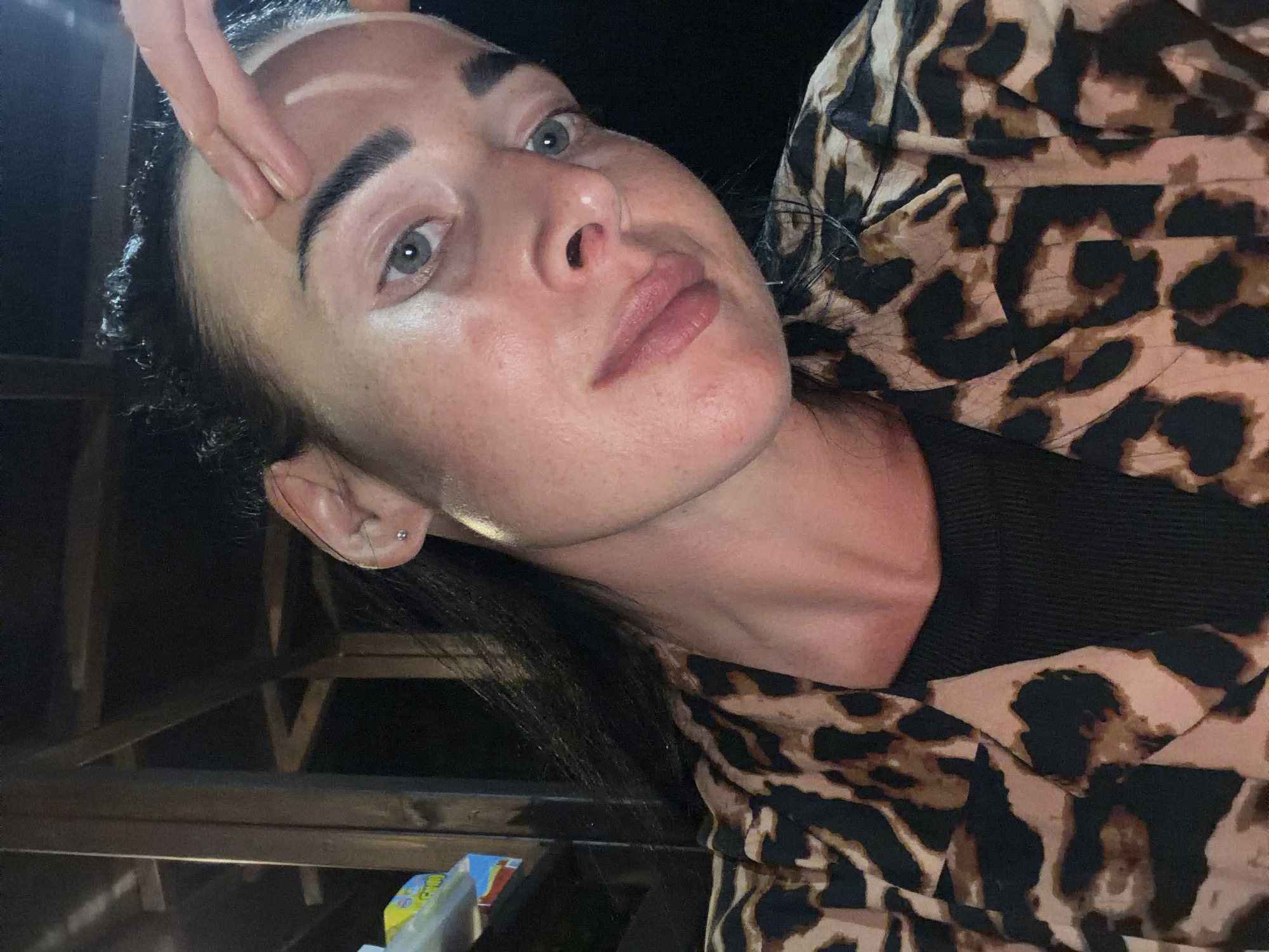 An outdoor, close-up, head-and-shoulders selfie is taken at night by a woman in Sochi, Russia. The woman has dark hair, fair skin, and light blue eyes. Her hand is touching her forehead. She is wearing a black top and a leopard-print shirt. The background appears to be a dark outdoor area with wooden structures, possibly part of a building or pavilion.
