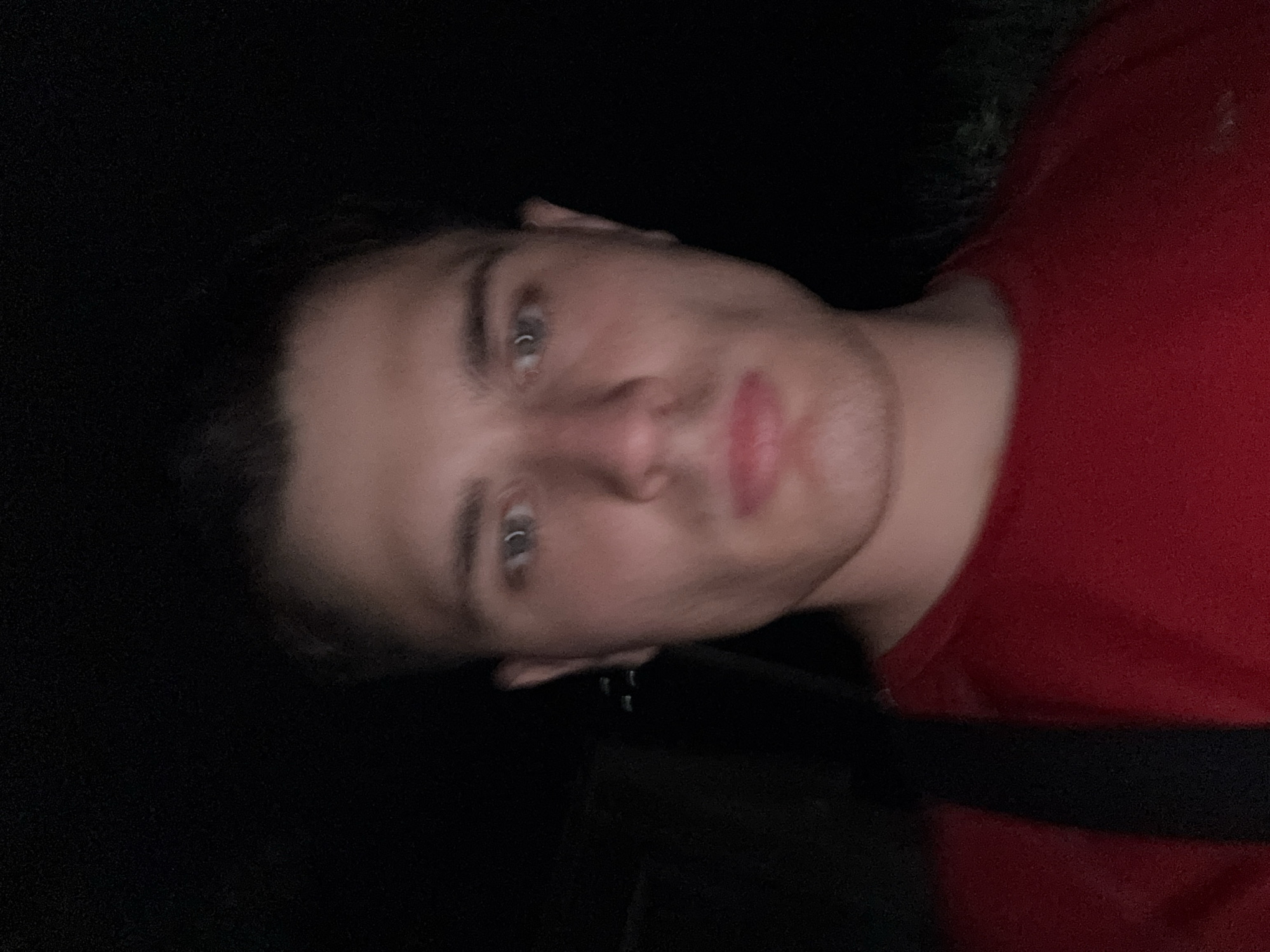 A young man with light skin, short brown hair, and light blue eyes is pictured in a close-up, dimly lit selfie. He is wearing a red t-shirt and a black strap is visible over his left shoulder. The background is dark, with some indistinct greenery visible on the right. There is no discernible location in the city of Stara Zagora, Bulgaria, or any other specific geographic information evident in the image. The man appears to be looking directly at the camera with a neutral expression.