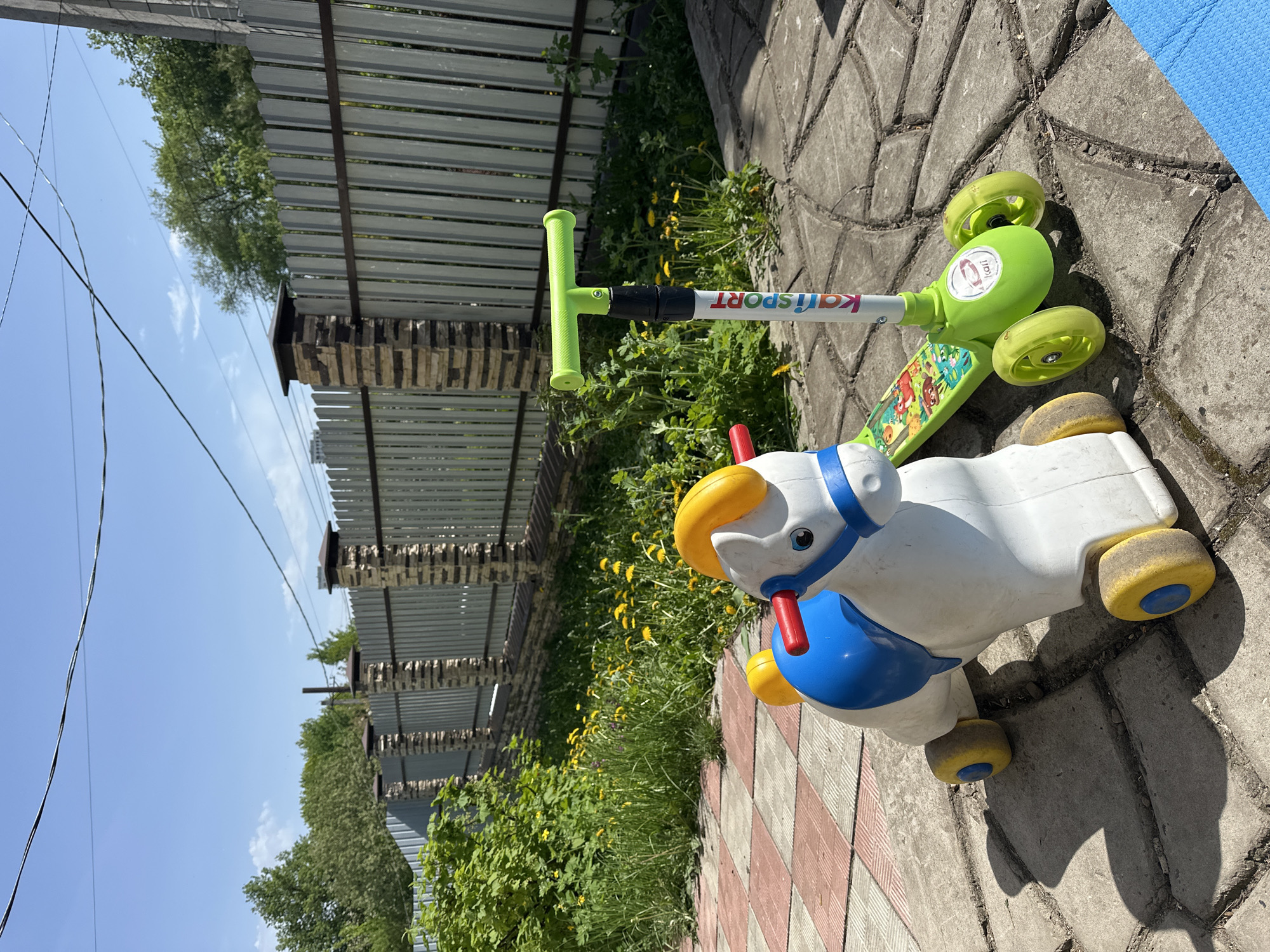 The image shows a children's scooter and a ride-on toy horse on a paved surface, possibly a patio or walkway. The scooter is green and white with the word "Kari Sport" visible on the handle. The horse is predominantly white with yellow and blue accents. In the background, there is a fence and greenery, including dandelions. The location is outdoors, with a blue sky visible.
The image does not contain any explicit clues indicating that the location is Nizhny Novgorod, Russia.