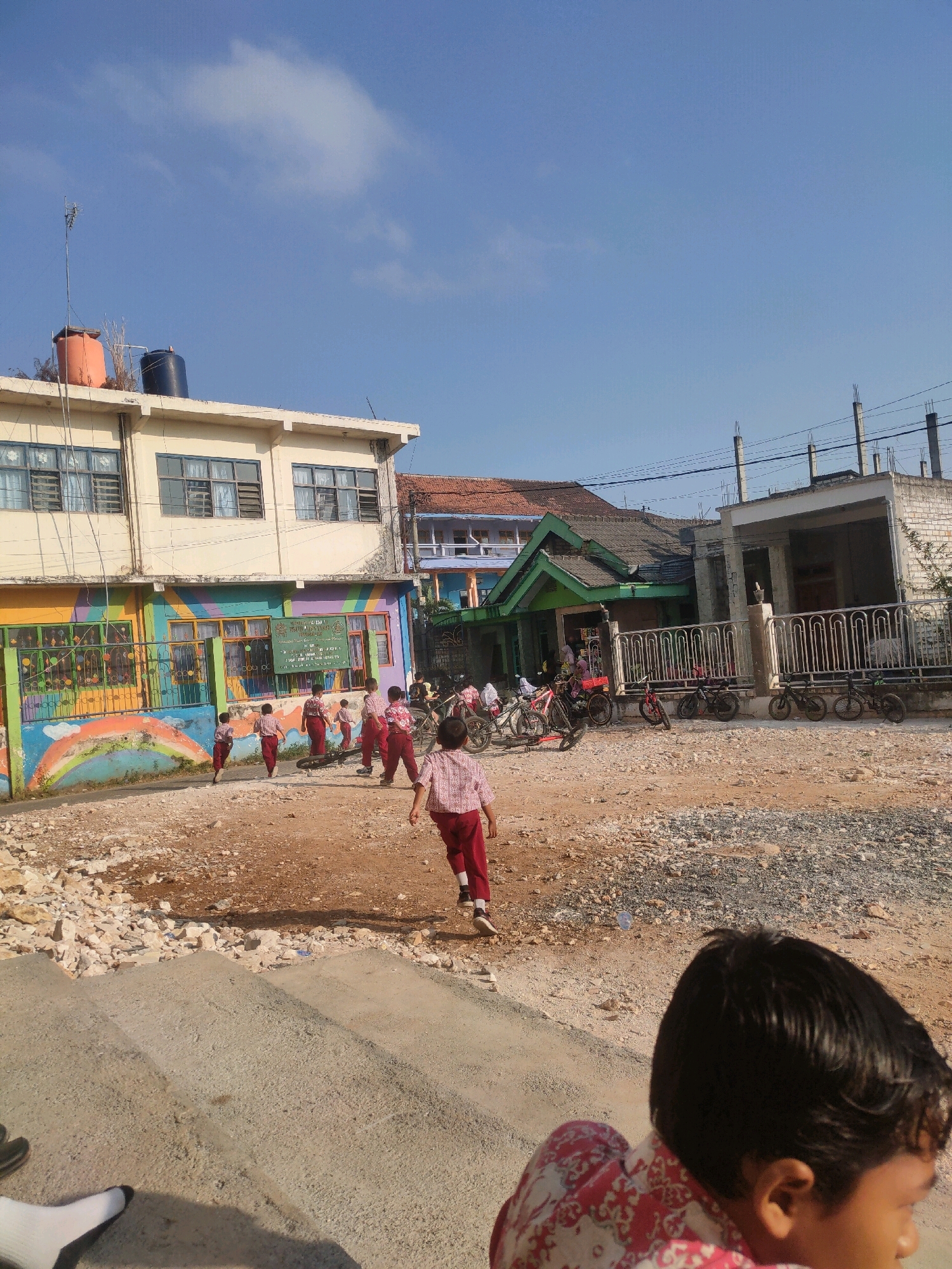 This image captures a scene outside a school in the city of Payaman, Indonesia, on a clear day. A group of young students, dressed in school uniforms of red pants and white shirts, are visible. Some are walking away from the camera, while others are in the foreground, also moving away. The school building on the left is a two-story structure with yellow walls and visible windows, featuring a colorful mural of a rainbow and various shapes on its lower level. In front of the building, a metal fence is present, and beyond it, parked bicycles are lined up. The ground is rough and unpaved, with scattered rocks and dirt. The background shows more buildings and utility poles. The overall impression is one of an active school environment with children engaged in activities.