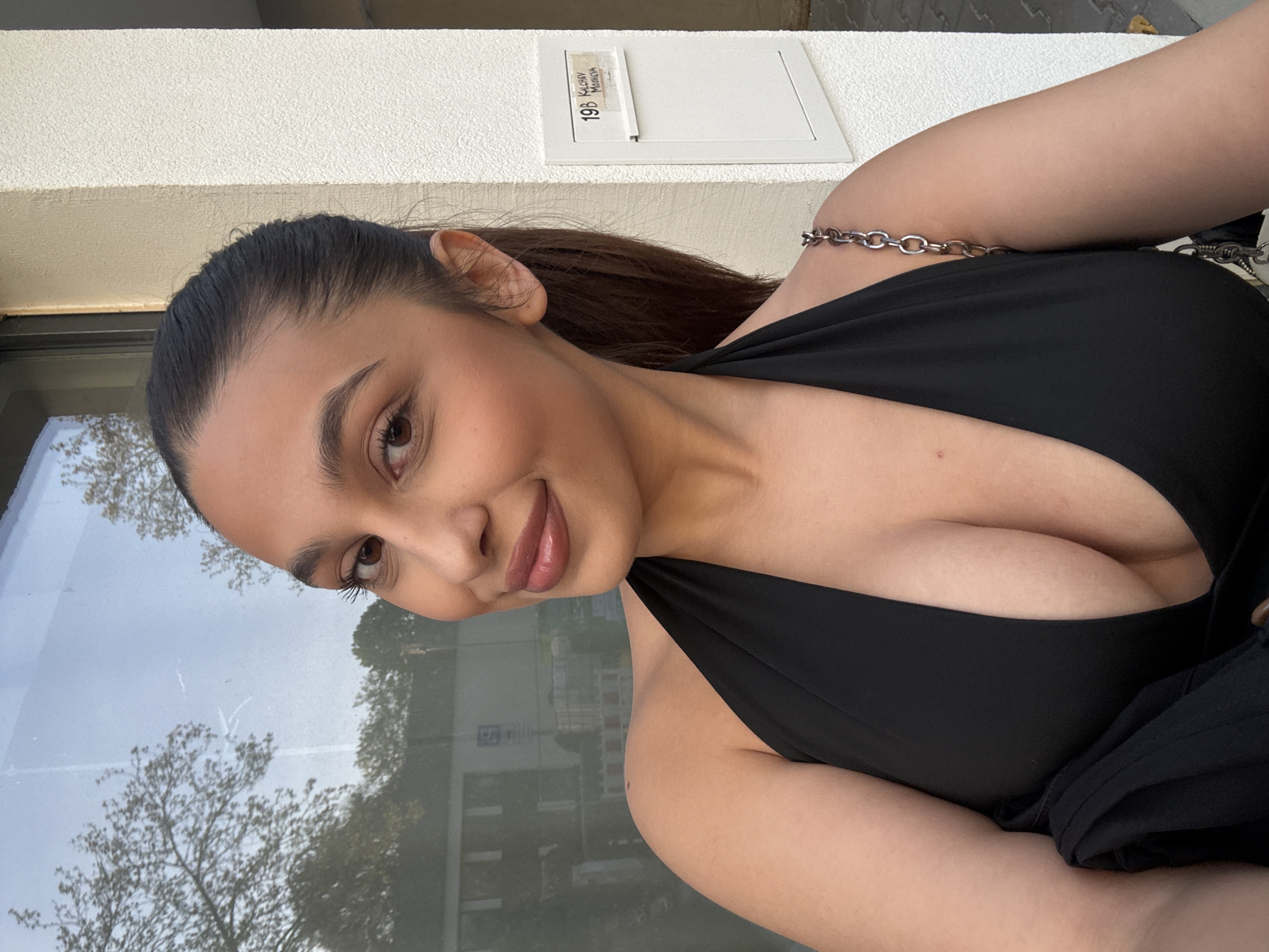A young woman is posing for a photo outdoors. She has dark hair pulled back from her face, with brown eyes and lips that appear to be glossy. She is wearing a black top with a low neckline, and a silver chain strap is visible on her shoulder.
The background includes a white wall with a small panel that has the address "19B Kalchov Mönchen" printed on it. There is also a window reflecting trees and buildings. The location is in Mönchengladbach, Germany, based on the address on the panel.