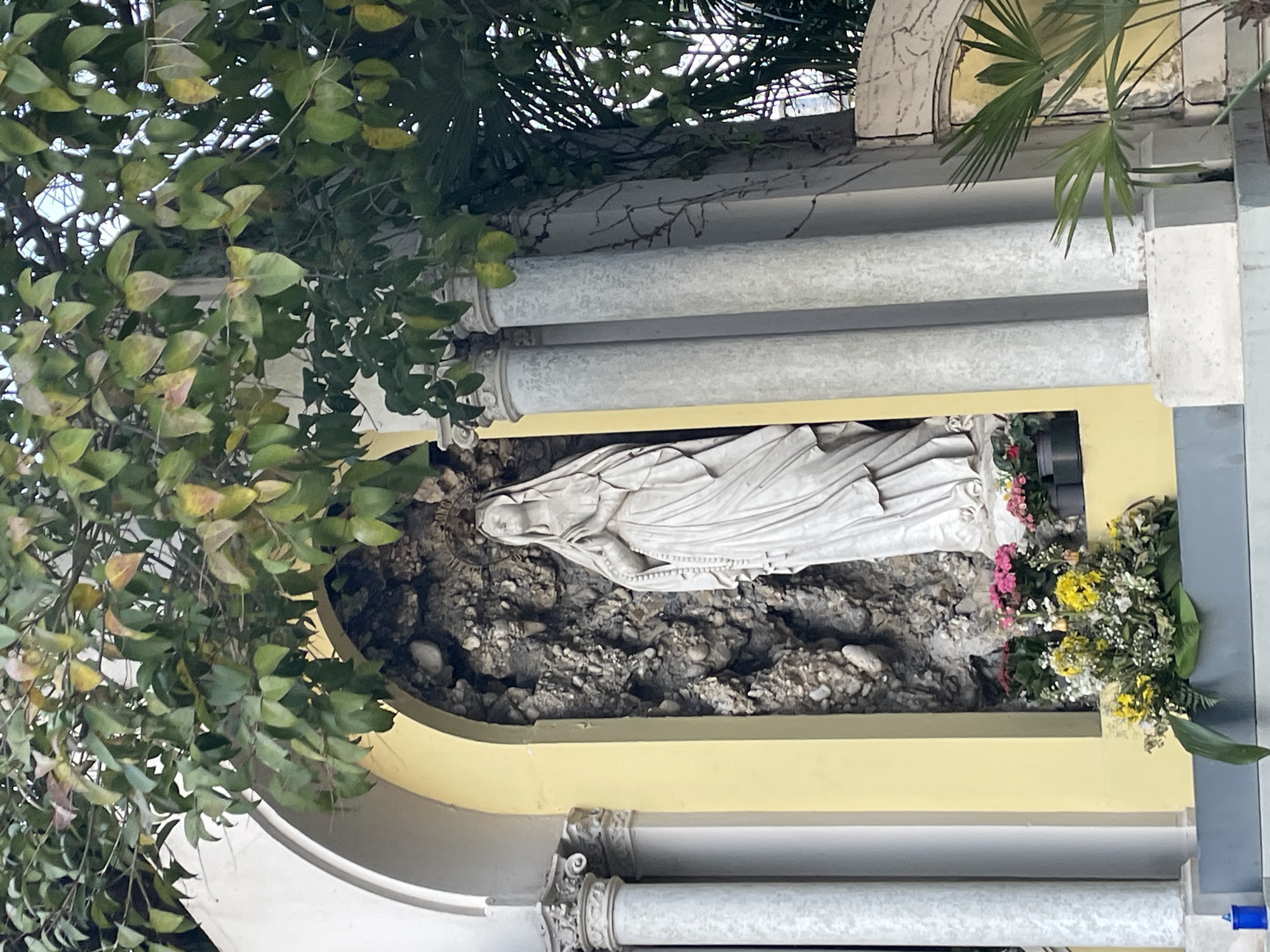 A low-angle shot captures a statue of the Virgin Mary within a recessed structure, adorned with flowers and foliage, in what appears to be an outdoor setting. The statue is white, depicted lying down, possibly in repose. It is situated within a grotto-like alcove filled with dark, textured stones, creating a naturalistic backdrop. To the statue's right, a colorful arrangement of pink, yellow, and white flowers adds a vibrant contrast to the stone setting.
The alcove is framed by a pale yellow structure that is part of a larger architectural element featuring stacked, decorative white beams. Lush green foliage, including vine-like plants with rounded leaves, drapes over the left side of the structure, softening its lines and providing a sense of organic growth.
The image is consistent with a religious shrine or memorial, potentially in a garden or public space. It is plausibly located in Case / Scjasis, Italy, given the architectural style and the subject matter.