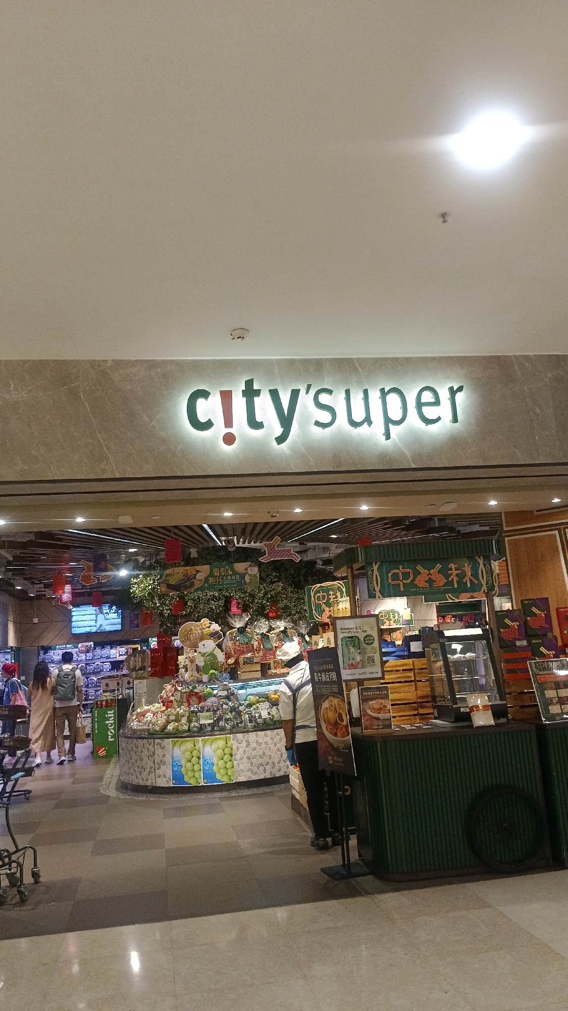 An exterior shot shows the brightly lit entrance of a City'Super store in Hong Kong, China. The store's name is prominently displayed in large, illuminated green lettering, with "city's" in a stylized font and "super" in a simpler font. The entrance features a modern, sleek design with marble-like accents and overhead lighting.
Inside the store, a display of fresh produce, possibly fruits and vegetables, is visible, arranged in a circular layout. Decorative elements, including red lanterns and a leafy backdrop, suggest a festive or seasonal theme, possibly related to a Chinese festival like the Mid-Autumn Festival, with rabbit motifs appearing. Several customers are seen browsing the merchandise. One person, facing away from the camera, is wearing a white shirt and dark pants, possibly a store employee. Another customer, also facing away, wears a beige dress and carries a shopping bag. Two other individuals are partially visible further down.
The store appears to be well-stocked with various food items, with displays of packaged goods and chilled items in refrigerated cases. A sign in Chinese characters is visible near the entrance, along with promotional materials and what looks like a QR code for a downloadable app. The flooring is a polished, tiled surface in shades of gray and beige, reflecting the overhead lighting. The overall atmosphere is that of a bustling, upscale supermarket.