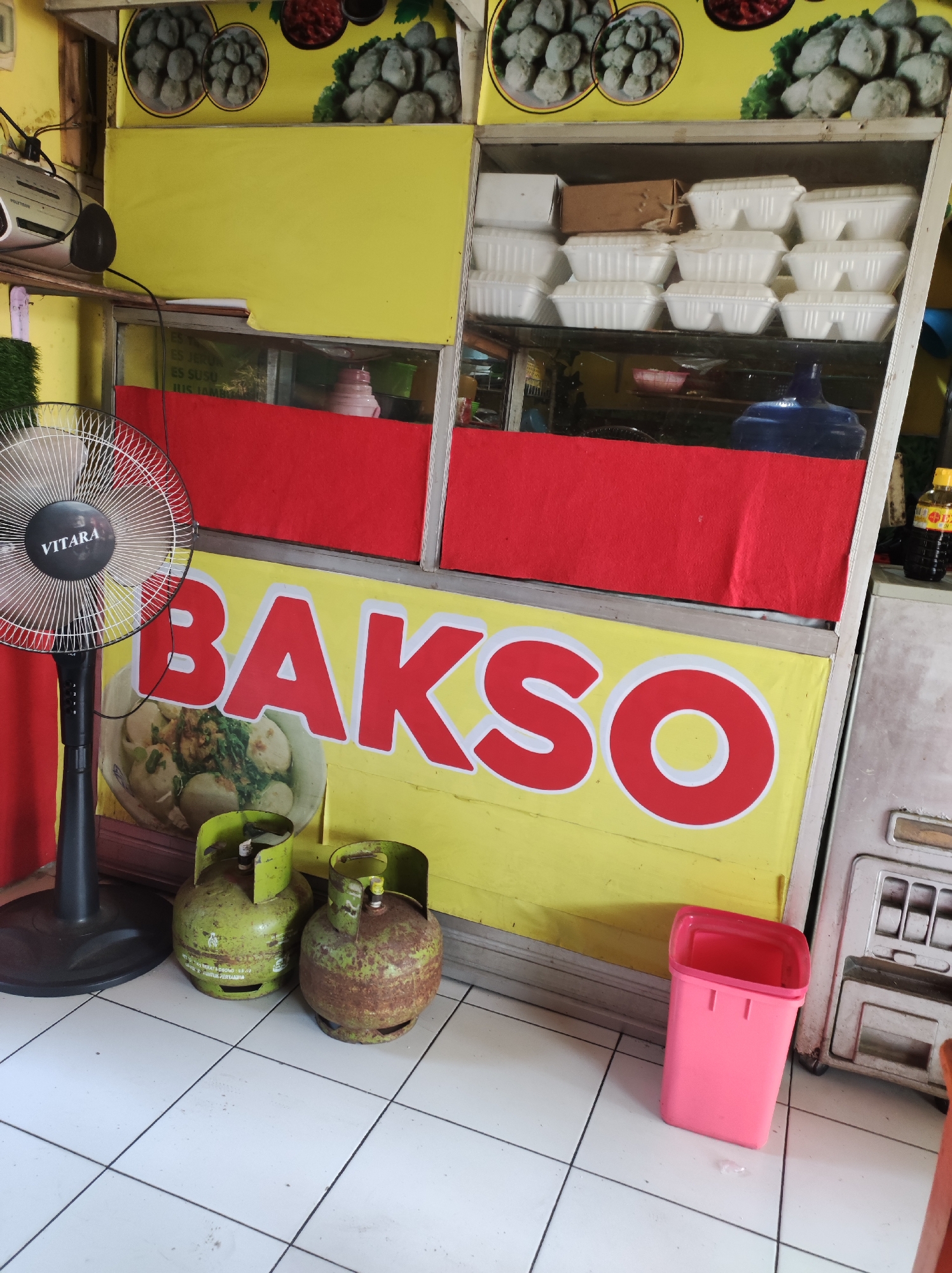 An outdoor food stall in Blitar, Indonesia, features a prominent yellow banner with the word "BAKSO" in large red letters. To the left, a black oscillating fan with the "VITARA" brand is positioned next to two green gas cylinders. Behind the banner, stacked white styrofoam containers are visible on shelves. A red fabric panel separates the upper and lower sections of the stall. A small pink trash bin sits to the right of the gas cylinders. The stall is set against a tiled floor.