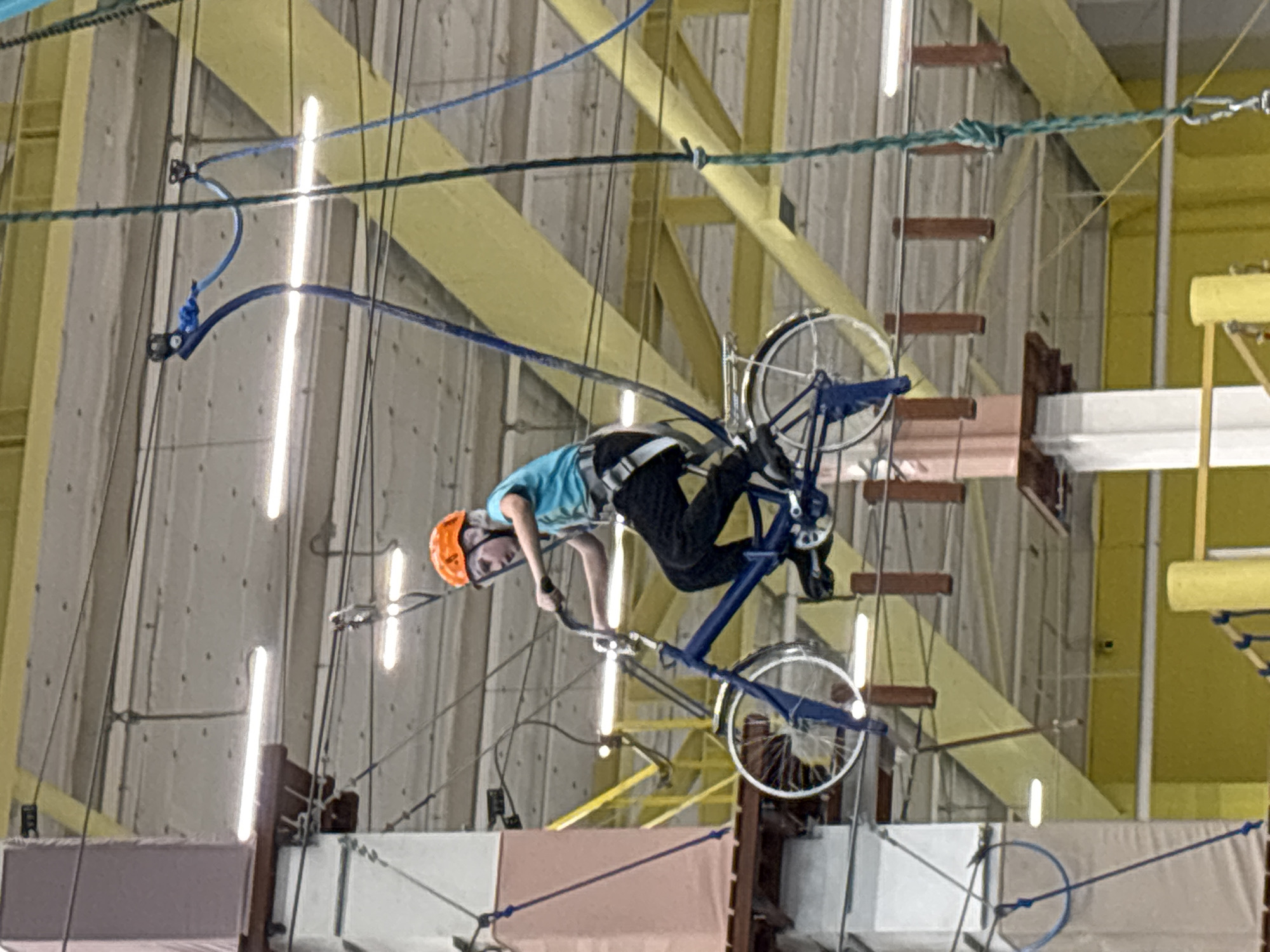 A young individual, identified as a pre-teen or teenager, is depicted engaged in an aerial adventure activity within an indoor high ropes course, likely located at Skala Perm, an adventure park in Perm, Russia. The person is situated on a blue bicycle, which is suspended approximately 5-7 meters above the ground, appearing to traverse or balance on an unseen narrow track or wire. The bicycle features white fenders and silver spokes.
The individual wears an orange safety helmet, a light blue short-sleeved t-shirt, and dark trousers. A grey safety harness is visible around their waist and thighs, with blue safety lines extending upwards from the harness to an overhead cable system, ensuring fall protection. Their hands are gripping the bicycle handlebars, and their feet are on the pedals, indicating active participation.
The surrounding environment consists of a complex network of high ropes course infrastructure. Wooden platforms and walkways, some with light pink or white padded coverings, are visible to the left and below the subject, supported by white and brown vertical pillars. Numerous ropes (blue and green) and cables crisscross the upper space, forming various challenge elements. Overhead, a light-colored, possibly concrete, ceiling is illuminated by several long fluorescent tube lights. Yellow structural beams are prominent across the ceiling and in the background, suggesting a large, industrial-style building. Yellow walls are also visible in the lower background.