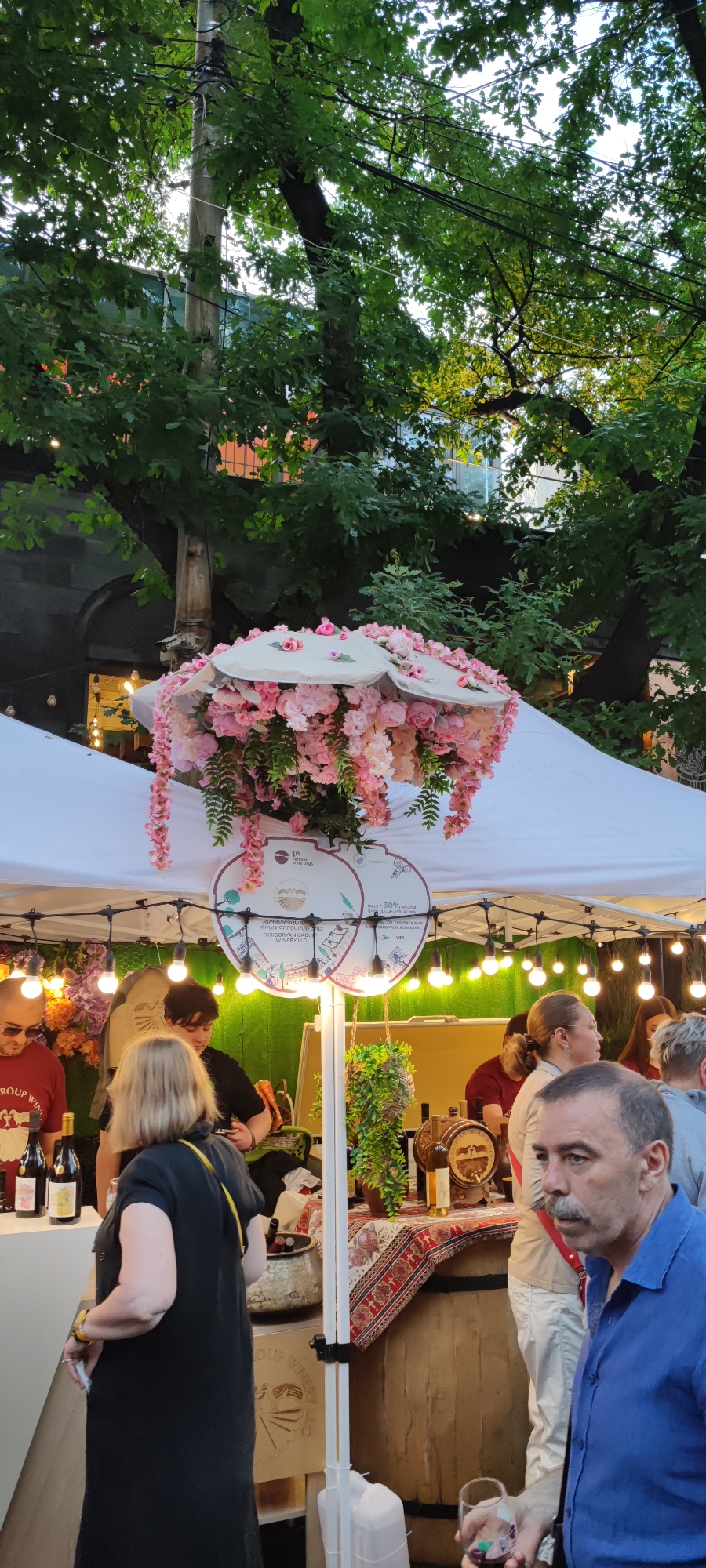 This image captures an outdoor event, likely a wine festival, in Yerevan, Armenia. The scene is illuminated by string lights, suggesting it is evening or dusk.
In the foreground, a white canopy structure provides shelter. Adorning the underside of the canopy are two circular, umbrella-like fixtures heavily decorated with artificial pink flowers, resembling wisteria and other blossoms, with some green foliage. Below these decorations, two approximately circular signs hang from a central pole. The left sign displays "10 Wine Days" and a logo, while the right sign prominently features "ԳՐԻԳՈՐՅԱՆ ԳՐՈՒՊ ԳԻՆԵԳՈՐԾԱՐԱՆ" (Grigoryan Group Winery LLC) in both Armenian and English, along with "30% cashback with ARDSHINBANK," and Visa and Mastercard logos. String lights are visible along the canopy's framework.
Several individuals are present around a vendor stall. On the far right, a man with a mustache, wearing a blue short-sleeved collared shirt, holds a small wine glass containing a red liquid. He looks slightly to his left. Behind him and to his left, other people are partially visible, including a woman with fair hair and a white top. In the mid-ground, a woman with blonde hair, wearing a black dress and a yellow strap across her shoulder, stands with her back partially to the viewer, facing the stall. To her left, a man in a red t-shirt with sunglasses pushed onto his head stands next to a display of wine bottles on a white surface.
The stall features a wooden barrel serving as a counter, adorned with a patterned red and white textile. On this counter, a small wooden wine barrel with a tap, additional wine bottles, and potted greenery are displayed. A large white plastic jerrycan rests on the ground near the man in the blue shirt.
The background is filled with dense green tree foliage, through which a tall utility pole with numerous wires is visible. Portions of urban buildings can be glimpsed beyond the trees. A security camera is mounted on a building structure visible through the trees on the upper left side of the image.