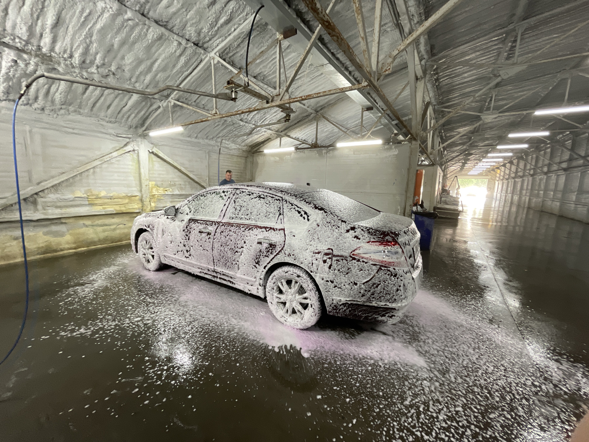 This media file captures a car being washed inside a tunnel-like car wash bay, likely in Moscow, Russia. A dark-colored sedan, possibly maroon or dark red, is positioned centrally in the bay, facing away from the camera, and is completely coated in a thick layer of white car wash foam. Some of the foam on the wet, dark floor, particularly near the car's wheels, has a distinct pinkish tint.
In the background, on the left side of the car, a man in a blue short-sleeved shirt stands, appearing to oversee or operate the car wash. A blue hose is visible on the left, extending towards the front of the car.
The interior of the car wash facility features a high, vaulted ceiling and upper walls covered in white or light gray spray foam insulation, giving it a textured appearance. Exposed metal trusses and structural beams support the roof. Numerous bright, rectangular LED or fluorescent light fixtures are mounted along the ceiling, providing strong illumination. The floor is dark, wet, and reflective, covered with scattered soap suds from the washing process. At the far end of the tunnel-like structure, bright natural light indicates an exit to the outside, suggesting it is daytime. No discernible text is visible.