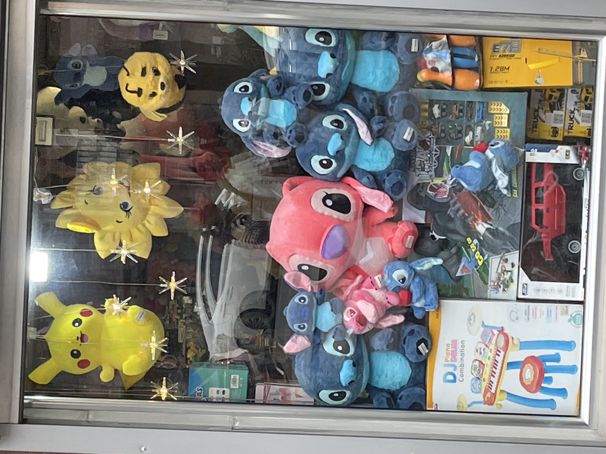 This image shows a storefront window display filled with various toys and plush items. On the left, there are plush toys shaped like Pikachu and sunflowers. The right side of the display has several plush toys of the character Stitch, including a pink version, along with boxes of toys like a DJ piano drum combination set, toy trucks, and other miscellaneous items. The scene appears to be in Yerevan, Armenia, but further details like exact location or store name aren't discernible. The items are densely packed within the window, giving the impression of a toy store or gift shop.