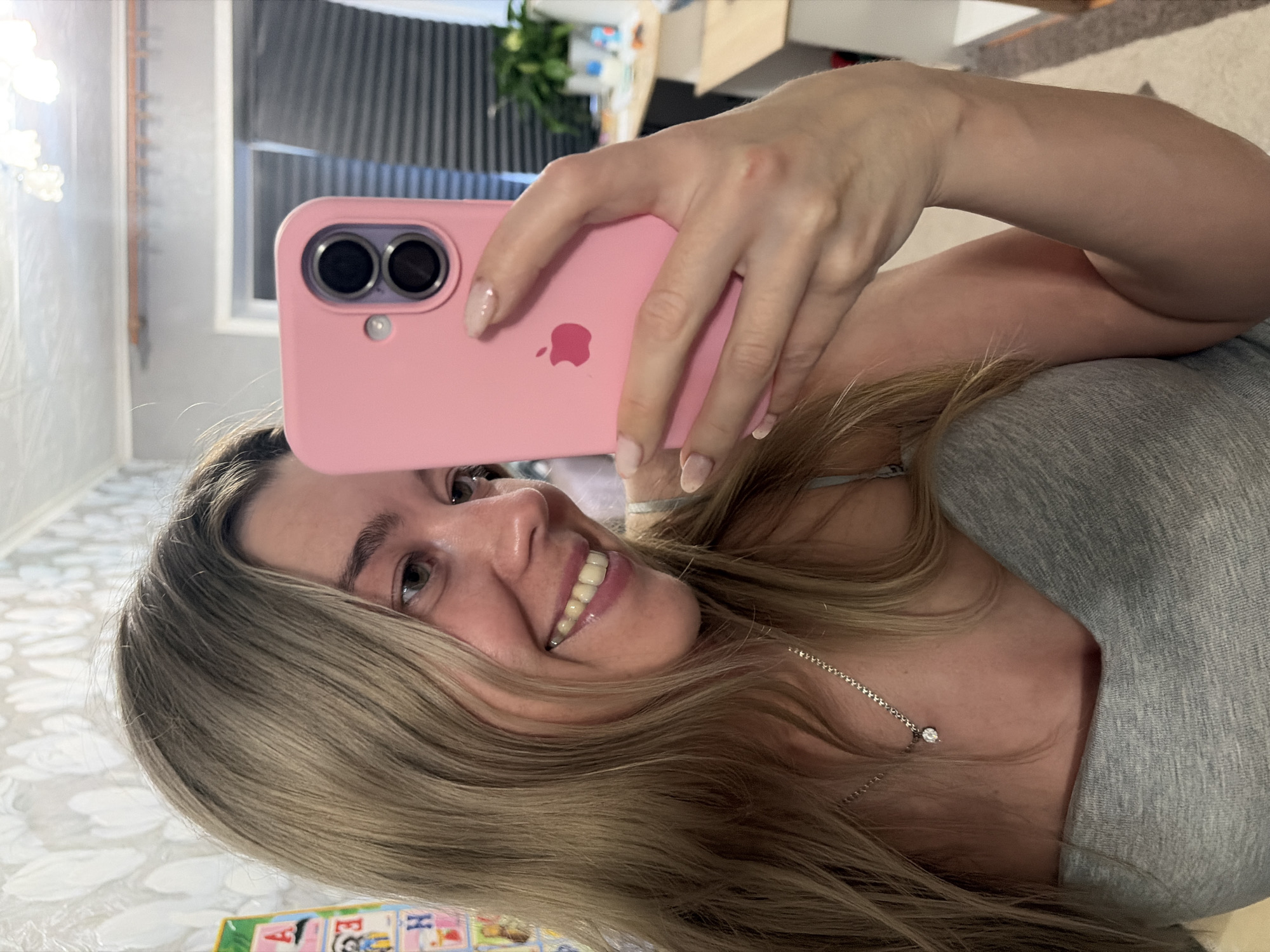 A young woman with blonde hair, wearing a gray tank top and a silver necklace with a diamond pendant, is taking a selfie in her home. She is smiling and holding a pink iPhone in her right hand, angled to capture her face and upper body. The background shows a wall with light-colored floral wallpaper, and a glimpse of a chandelier and a closet door. The setting appears to be an indoor residential environment, likely a bedroom or living area. The photograph was taken in Chaikovsky, Russia.