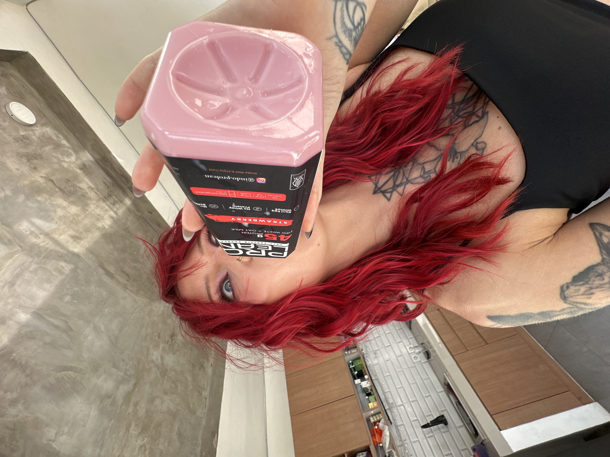 A woman with bright red hair and tattoos is drinking from a pink bottle of Prolean. She is wearing a black tank top and is in a kitchen with wooden cabinets and white tiled backsplash. The scene appears to be an apartment kitchen.