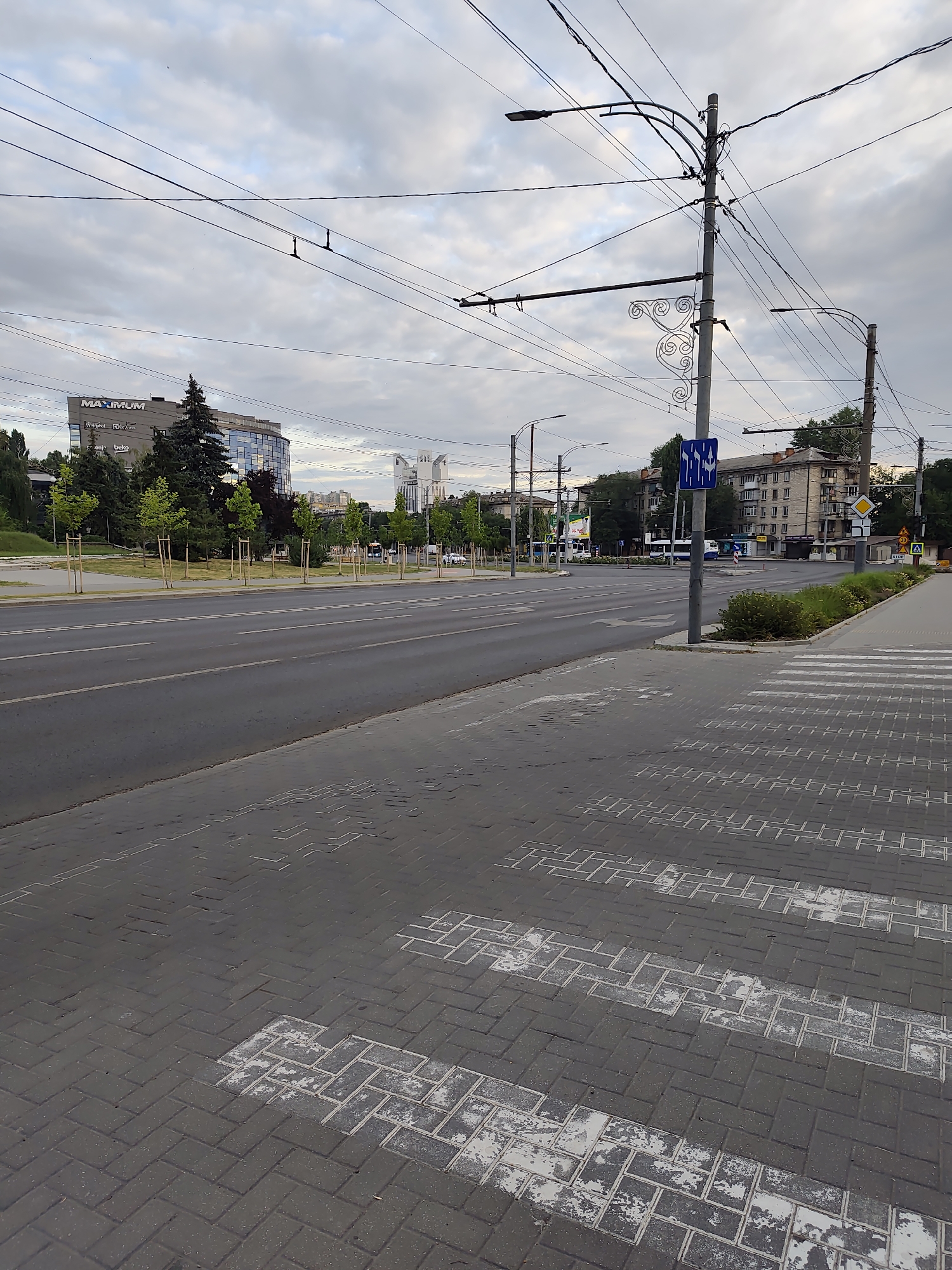 The image depicts a street scene in Chișinău, Moldova, likely near the intersection of Stefan cel Mare Boulevard and Ismail Street. The foreground is dominated by a paved pedestrian crossing with worn white markings. Beyond the crossing, a wide asphalt road stretches into the distance, with traffic lights and overhead power lines characteristic of a city environment. On the left side of the road, a modern, curved building with the "MAXIMUM" logo is visible, surrounded by young trees. Further down the street, residential buildings and more trees line the road. The sky is overcast, suggesting a cloudy day. There are no people or vehicles prominently featured in the image, giving it a sense of stillness. The overall impression is that of an urban streetscape under a subdued sky.