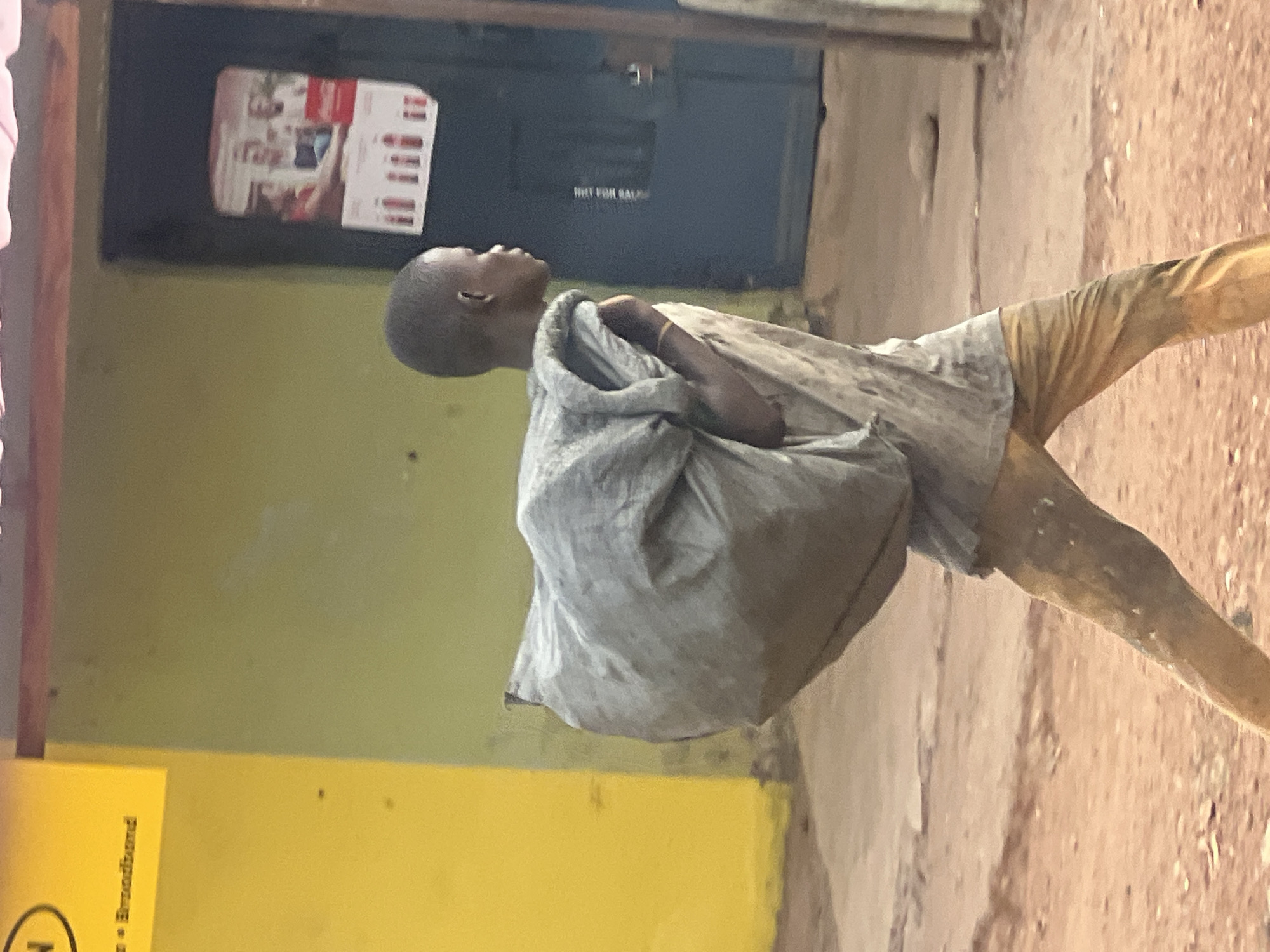 A young person walks in the city of Gura Top, Nigeria, carrying a large, light-colored sack over their shoulder. The individual wears light-colored, dirty clothing and has a shaved head. The background shows a building with yellow and green walls, and a dark door with a sign on it. The ground is a mixture of dirt and pavement.