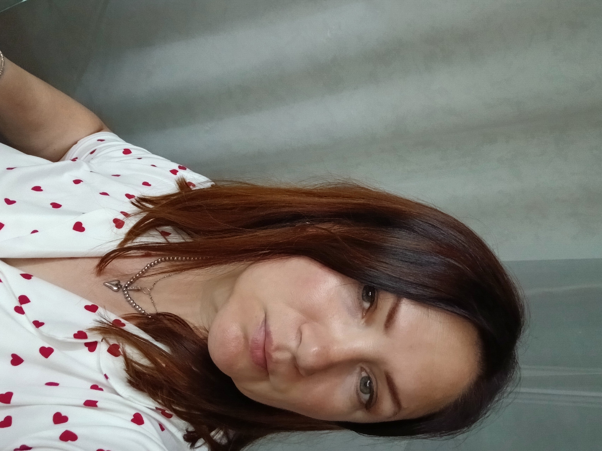 A medium shot portrait shows a woman with medium-length auburn hair, facing slightly to the right. She is wearing a white shirt with a pattern of red hearts and a silver necklace with a heart-shaped pendant. Her expression is neutral. The background is a textured gray wall. The photo appears to have been taken indoors.