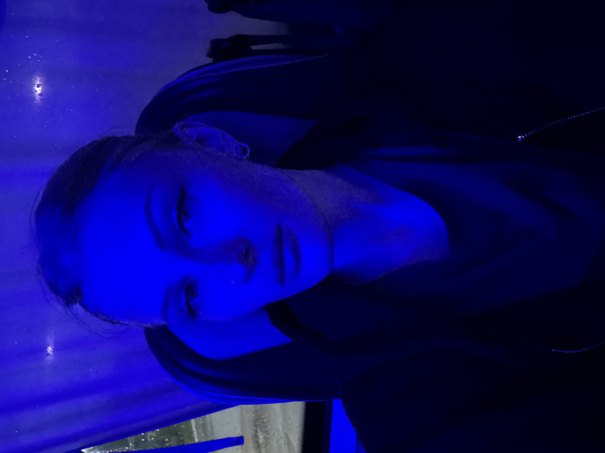A young woman with blonde hair pulled back in two ponytails poses for a selfie. She is illuminated by a bright blue light, which casts deep shadows on her face. She wears a black jacket over a dark shirt, and a circular, jeweled earring is visible in her left ear. The background is dark and out of focus, with some blurred shapes that might suggest seating or interior elements of a vehicle. The overall lighting and mood are dramatic and somewhat mysterious. The location is identified as the city of Kazan, Russia.