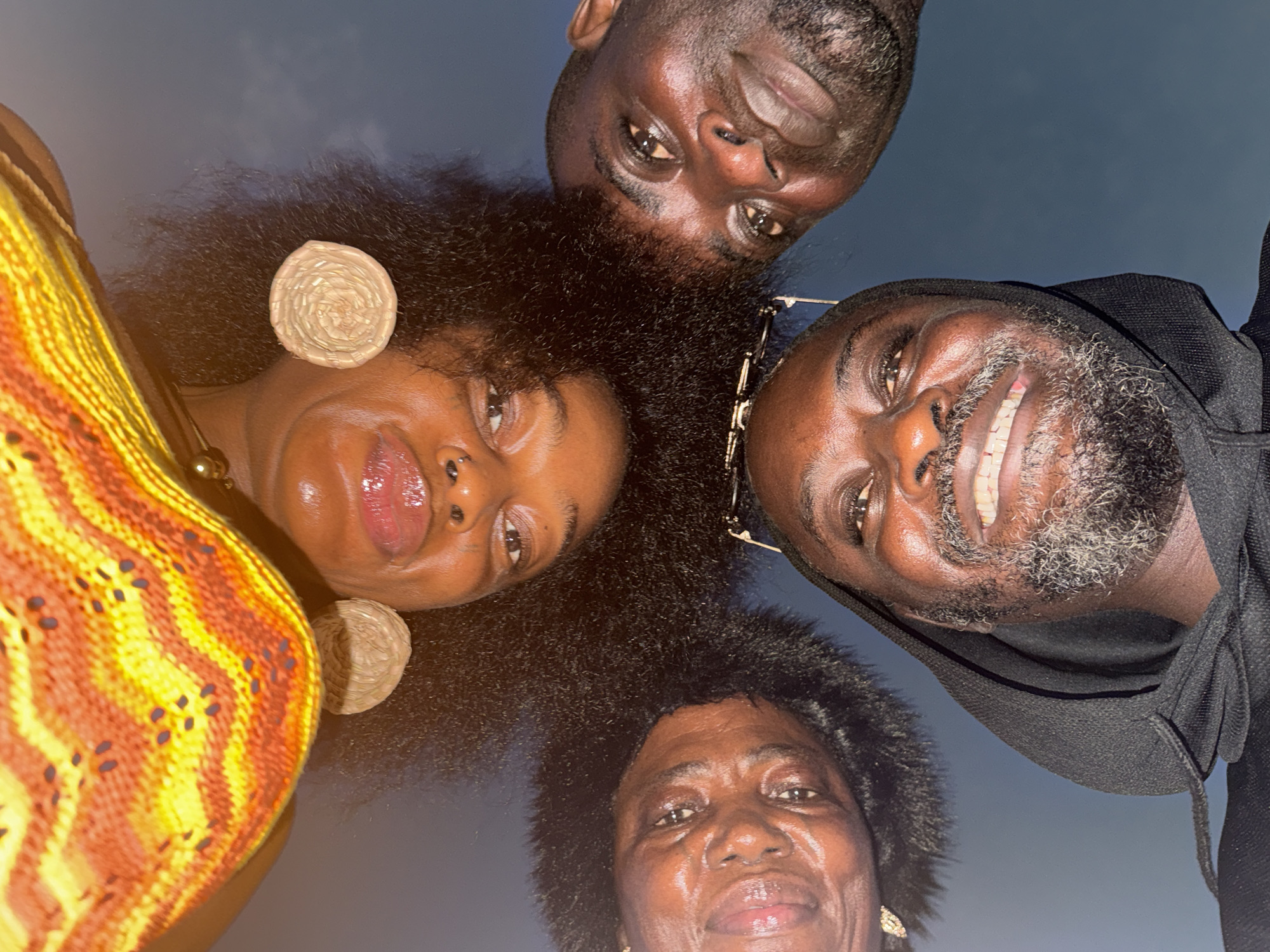 Four people, appearing to be African, are in close proximity, looking down at the camera. The background is a dark blue, possibly indicating nighttime or a dimly lit area.
The person on the left is wearing a yellow and orange patterned garment and large round earrings. The next person has dark, curly hair. The man on the right has glasses perched on top of his head and is wearing a black hooded sweatshirt. He also has a graying beard and is smiling. The final person at the bottom also has short, dark hair and gold earrings.
I am unable to locate a city called "Mopo Onijebu" in Nigeria. Therefore, I cannot verify the location.