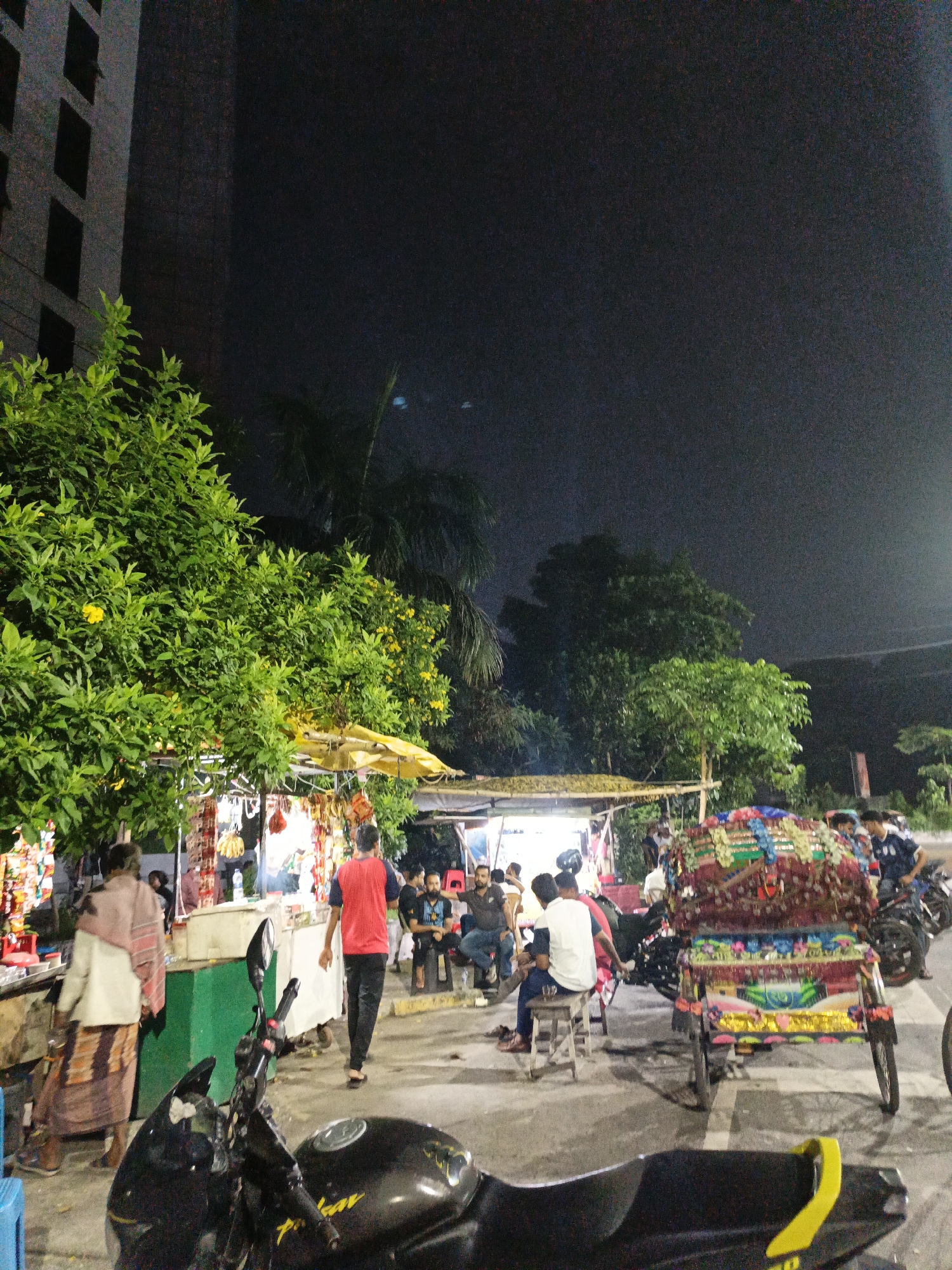 An outdoor, street-level, night-time scene captures a bustling market area with various vendors and patrons. A large, dark building is visible on the left, partially obscured by a vibrant green tree with yellow flowers. In the foreground, a black motorcycle is parked. To the right of the motorcycle, a decorated rickshaw sits prominently, its canopy adorned with colorful fabric and embellishments. Several people are gathered around market stalls, some seated on stools, while others are standing or walking. One stall displays a variety of goods, including what appear to be food items and packaged products. The ground is paved, and the overall atmosphere suggests a lively evening market.