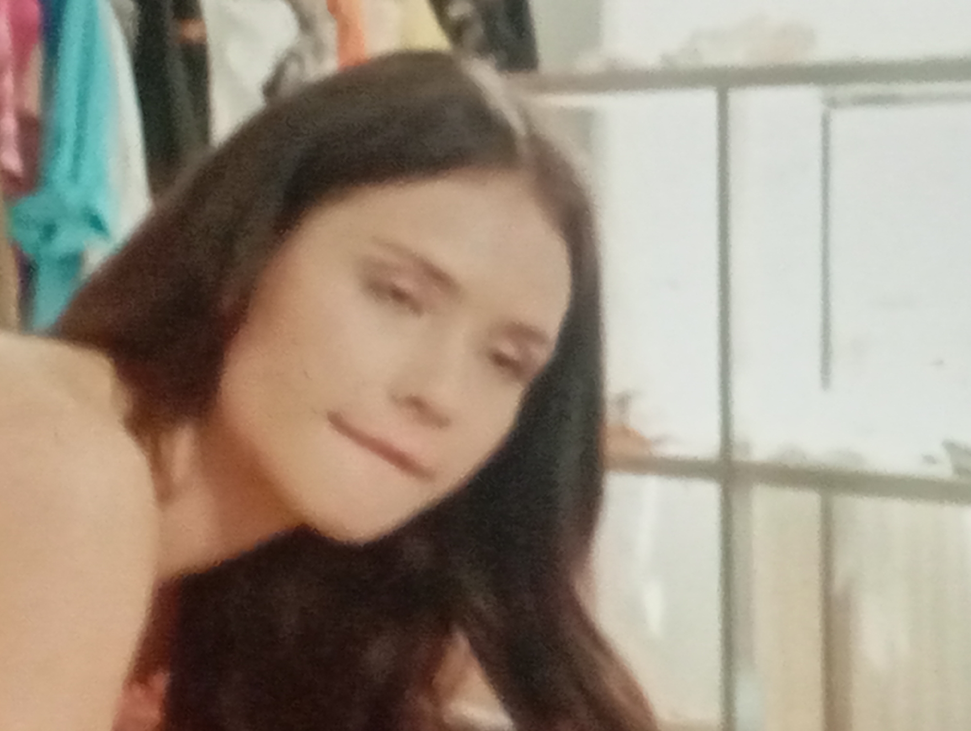 A young woman with long, dark hair is pictured in a shallow depth of field, her face angled away from the camera. The background suggests an indoor setting, possibly a closet or dressing area, with hanging clothes visible on the left and a shelf or divider on the right. The lighting is soft and diffused, creating a somewhat hazy atmosphere. The woman's expression is contemplative or slightly amused, with her lips parted. There is no indication of her location within Sofiivska Borshchahivka, Ukraine, nor any interaction with other individuals. The image is composed in a close-up shot, focusing on the woman's upper body and head.