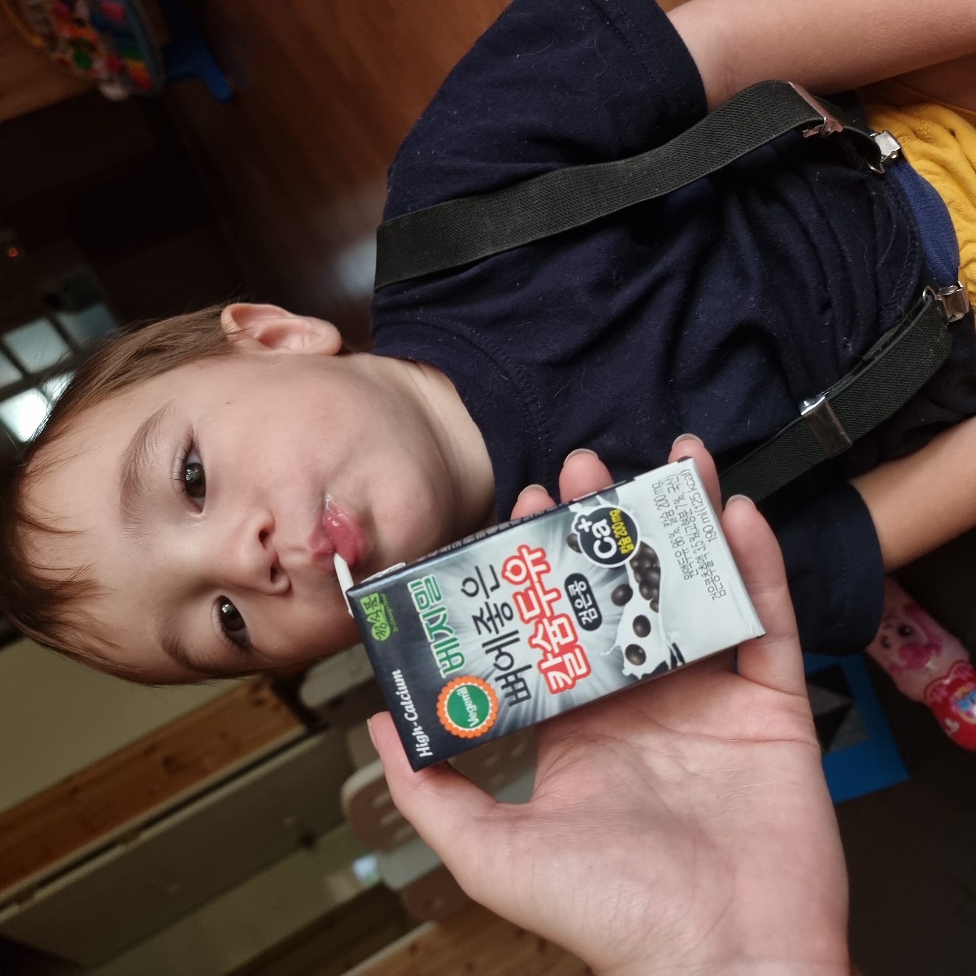 A close-up, eye-level shot shows a child drinking from a juice box held by a person's hand. The child, with brown hair and brown eyes, is wearing a navy blue shirt with suspenders and yellow shorts. The juice box, black and white with Korean writing and a picture of black beans, is labeled "High-Calcium" and "Vegemil." The background is blurred, showing a wooden floor and a toy. The scene appears to be indoors.