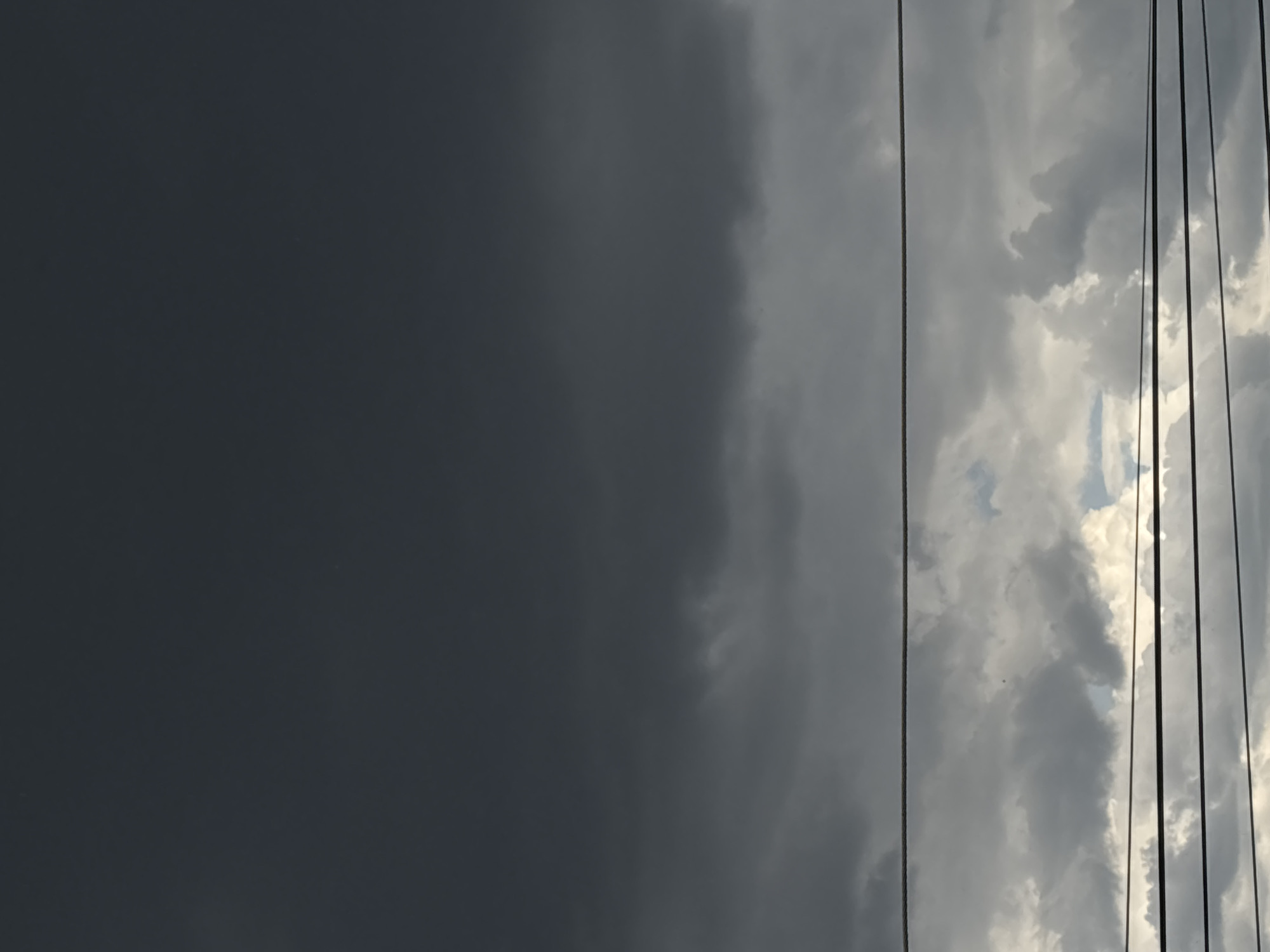 The image shows a sky divided into two distinct areas. The left side is dominated by a dark, ominous-looking sky, while the right side features lighter, cloudier skies with some glimpses of blue. Several vertical power lines are visible against the lighter clouds on the right. The location appears to be urban, likely a city where power lines run above ground. The city itself is unidentified, labeled as "Unknown, Unknown." No people, other subjects, actions, or interactions are apparent in the image. The environment is marked by contrasting weather conditions.