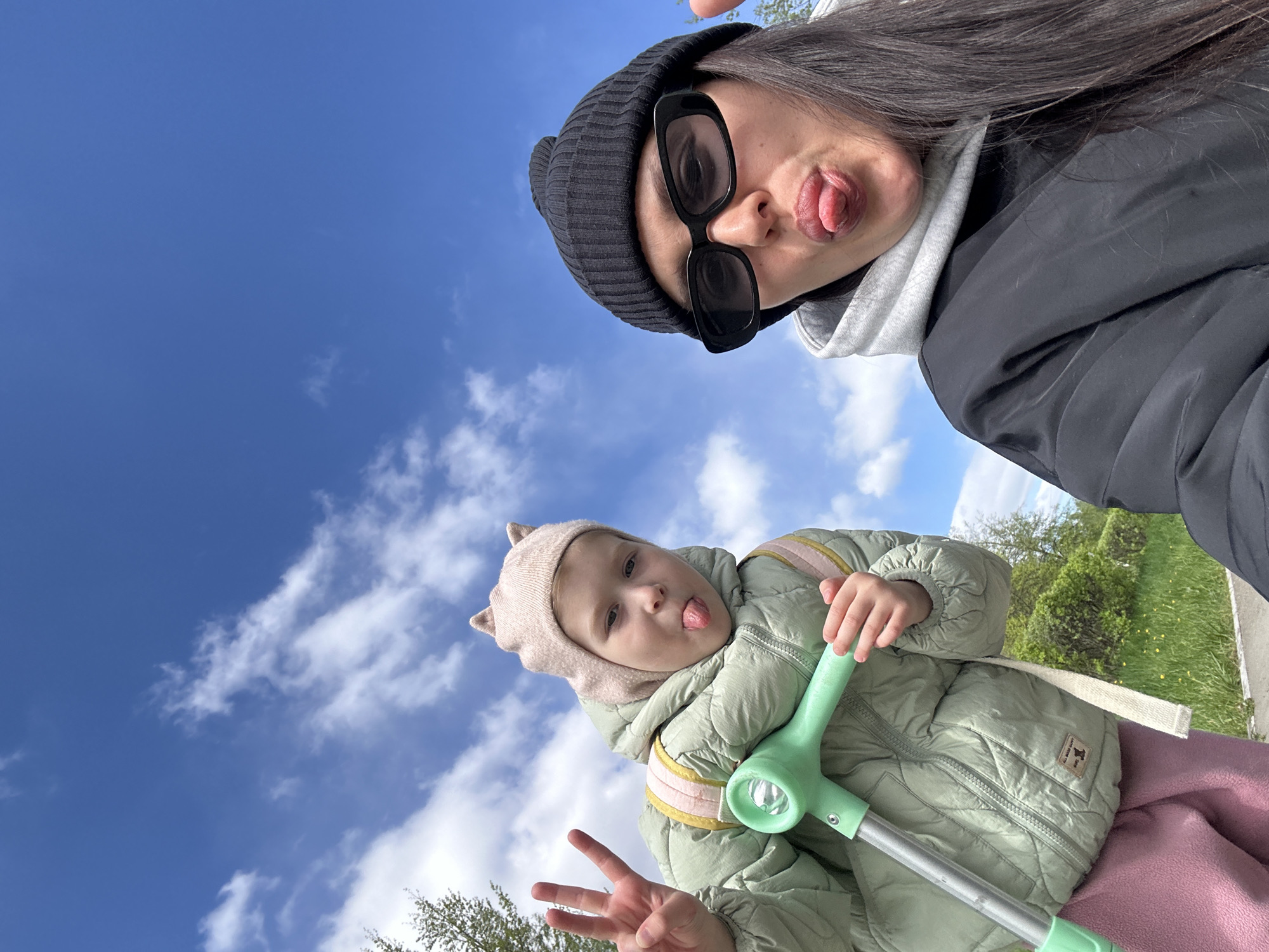 A selfie taken from a low angle features a woman and a young child outdoors against a backdrop of a bright blue sky with scattered white clouds. The woman, wearing a black beanie and black sunglasses, is at the top of the frame, sticking her tongue out at the camera. She is wearing a black jacket over a grey hooded sweatshirt. Below her, the child, clad in a pale green puffer jacket with a hooded beige hat adorned with small ears, also sticks out their tongue. The child is holding onto the handle of a light green scooter and makes a peace sign with their left hand. Some trees and greenery are visible in the background. This photo was taken in the city of Chaikovsky, Russia.