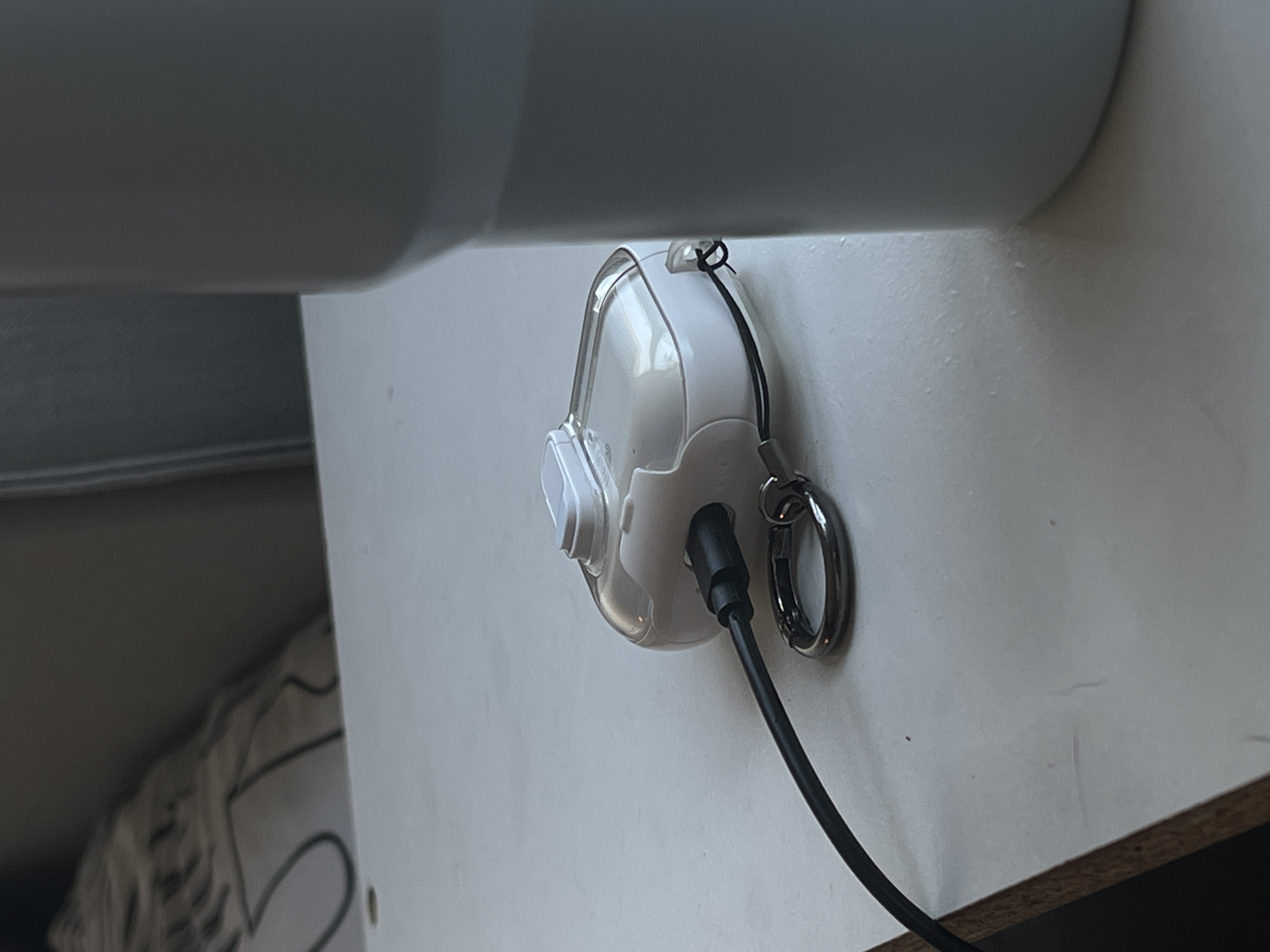 A clear plastic and white case, possibly for earbuds, is placed on a white surface. A black cable is plugged into the case, with a silver ring attached to the case via a black string. To the right of the case, a portion of a large, light gray tumbler is visible. The background shows a blurred pattern of a pillow and a dark surface, likely furniture. The scene is indoors and appears to be a tabletop in Genemuiden, Netherlands.