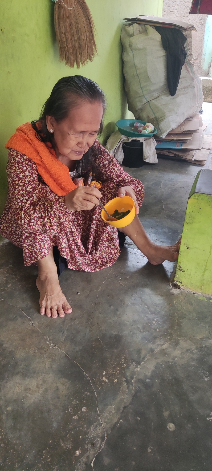 An elderly woman with dark hair and an orange scarf around her neck sits on a concrete floor in what appears to be a room in Karangwangun, Indonesia. She is wearing a floral print dress and is holding a small yellow bowl in her left hand, eating with a spoon from it. To her right, a green object, possibly a piece of furniture or wall, is visible. In the background, a large sack is leaning against a light green wall, and a broom is hanging on the wall above it. Several cardboard boxes are stacked to the right of the sack. A green basin containing small objects, possibly bottles and a towel, sits on the floor near the sack. The environment suggests a humble, possibly cluttered living space.