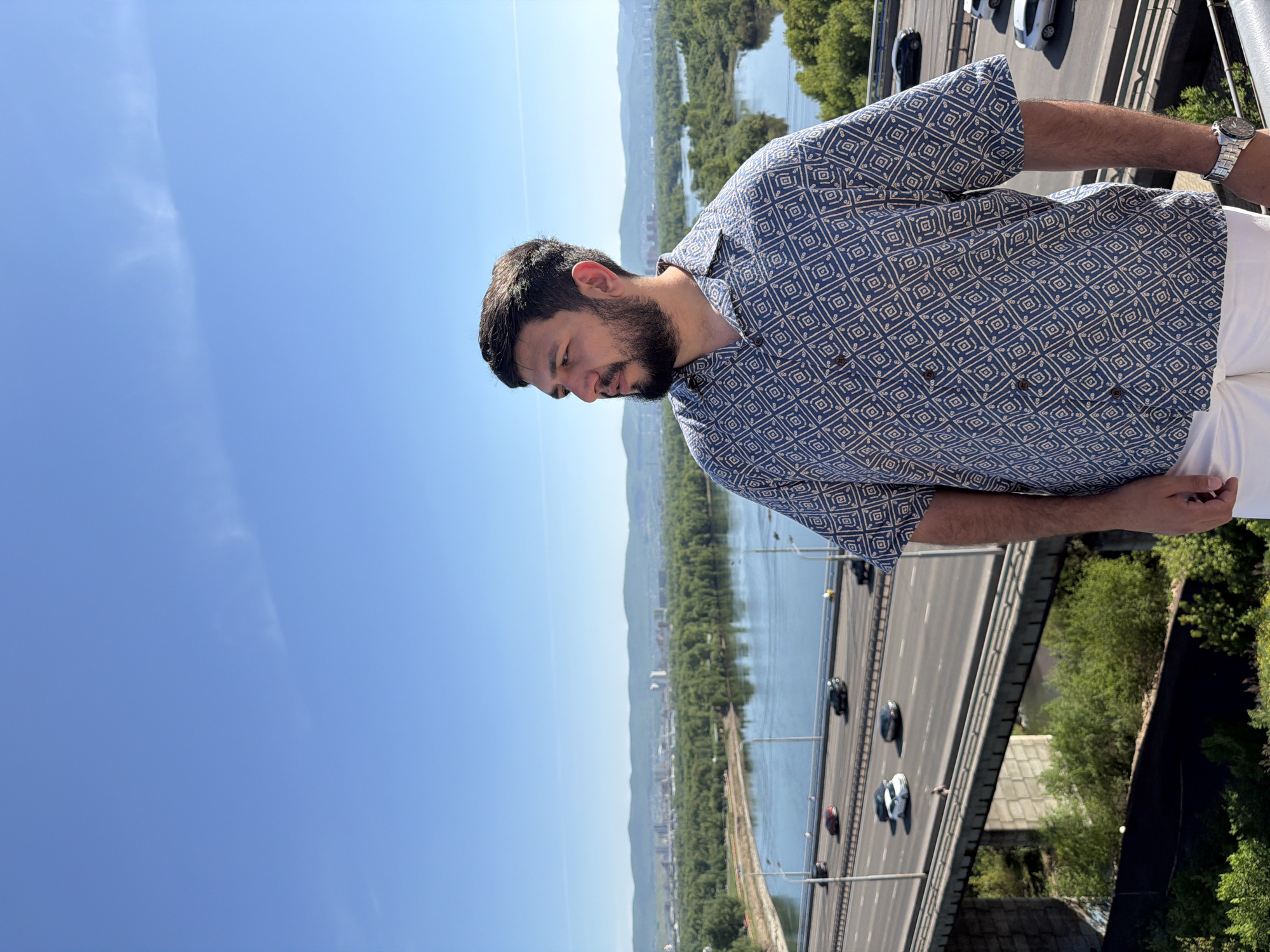 A male individual with dark hair, a full beard, and a mustache stands on a high vantage point, looking leftward in profile. He is wearing a short-sleeved, collared button-up shirt with a blue and off-white geometric pattern, and white trousers. A silver-toned watch is visible on his left wrist. Behind him, a panoramic view of the Yenisei River in Krasnoyarsk, Russia, unfolds under a clear blue sky with minimal wispy clouds.
In the midground, a multi-lane highway bridge spans the wide river. Several vehicles, including cars of various colors (red, black, white, silver), are visible on the bridge, moving in both directions. Below the main highway, a narrower bridge or structure is also present. The river's banks and several islands are densely covered with green trees and foliage. In the far background, the cityscape of Krasnoyarsk is visible on the opposite bank, backed by a range of distant green hills or mountains. The scene is brightly lit by sunlight, indicating daytime.