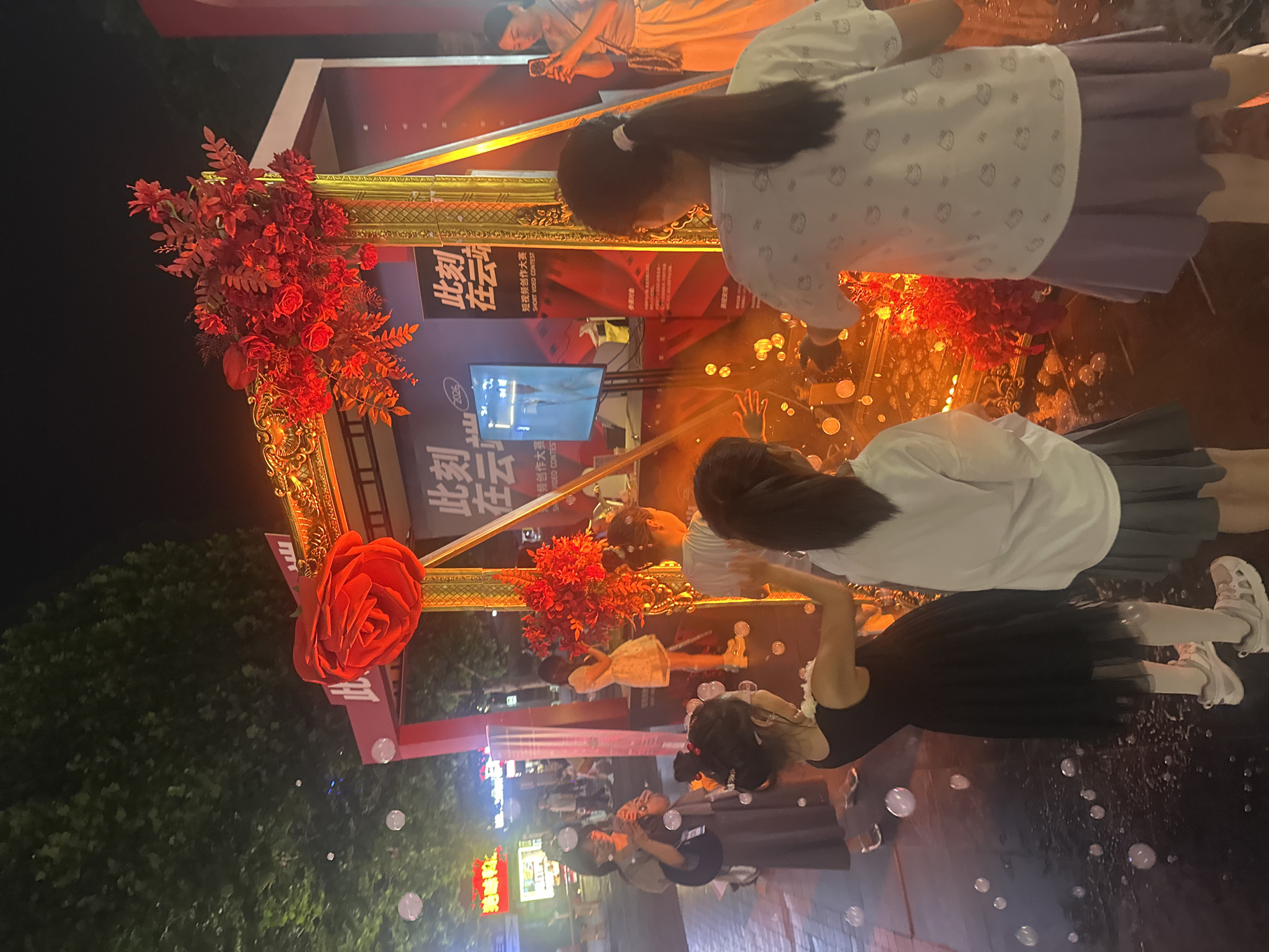 This image depicts a nighttime outdoor scene in the Chongqing High-tech Zone, China, centered around an illuminated, ornate golden frame structure. The frame is adorned with a large red rose at its upper left corner and multiple clusters of smaller red floral arrangements. Banners within and around the frame display Chinese characters, prominently featuring "此刻 在云端" (This Moment in the Cloud) and "短视频创作大赛" (Short Video Creation Contest), with "SHORT VIDEO CONTEST" also visible in English. A screen inside the frame shows a person.
Approximately six identifiable individuals are present, along with several blurred figures in the background. In the foreground, three young females face the display. A girl in a black dress, white tights, and athletic shoes actively interacts with numerous soap bubbles floating around and resting on the dark paved ground. Adjacent to her are two other young females; one wears a white T-shirt with repeating patterns and a purple skirt, and the other wears a white long-sleeved top and a grey skirt.
Two adult females are positioned on either side, observing or filming. On the left, a woman in a long brown dress holds a mobile phone, capturing the scene. On the far right, another woman in a beige top and light-colored pants uses a mobile phone to record the display. A third female in a light-colored dress is partially visible behind the right side of the frame. The scene is lit by the display's red and orange lighting, along with ambient streetlights. Dark trees and distant illuminated shop signs are visible in the background.
