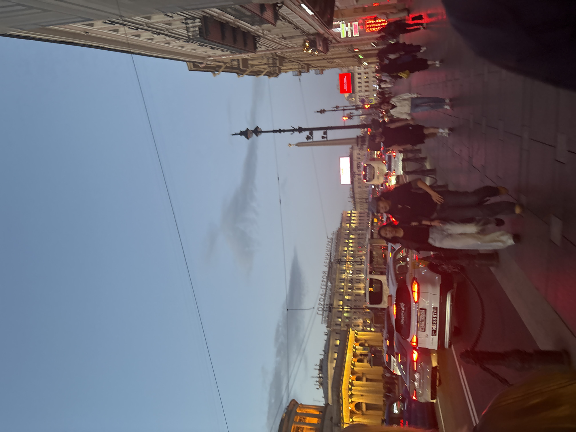 The image depicts a street scene in Saint Petersburg, Russia, likely during twilight. On the left, a grand building with illuminated columns is visible, possibly a historical structure. The center of the image is dominated by a street filled with cars, including a white SUV with a license plate that reads "A511EY 778". People are walking on the sidewalk on the right side of the street. Further down the street, an obelisk stands tall, a prominent landmark. The sky is a pale blue, with some clouds. The buildings on the right are multi-story structures with windows and balconies, suggesting an urban environment. The overall atmosphere is that of a busy city street in the evening.