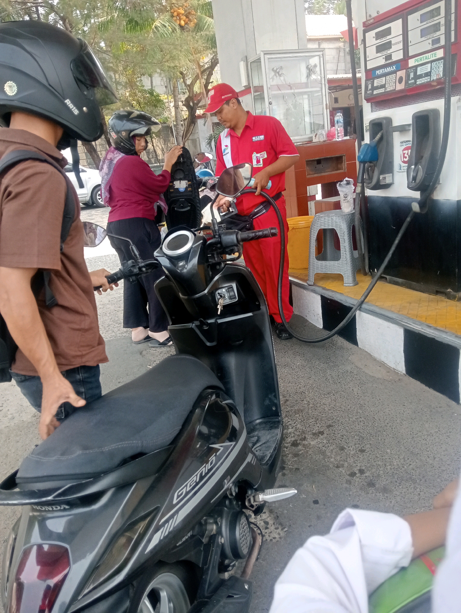 The image depicts a scene at a Pertamina gas station in Bandar Lampung, Indonesia. A gas attendant, wearing a red uniform and a red cap, is in the process of refueling a black motorcycle. The motorcycle is positioned near a gas pump, with its fuel cap open. A person in a brown shirt and jeans is standing to the left of the motorcycle, holding the handlebar. Another person wearing a maroon shirt and a black helmet is standing behind the motorcycle, holding the seat. A white car is visible in the background, along with a palm tree and a building that appears to be part of the gas station. The ground is paved with asphalt. The scene suggests a typical transaction of refueling a vehicle at a gas station.