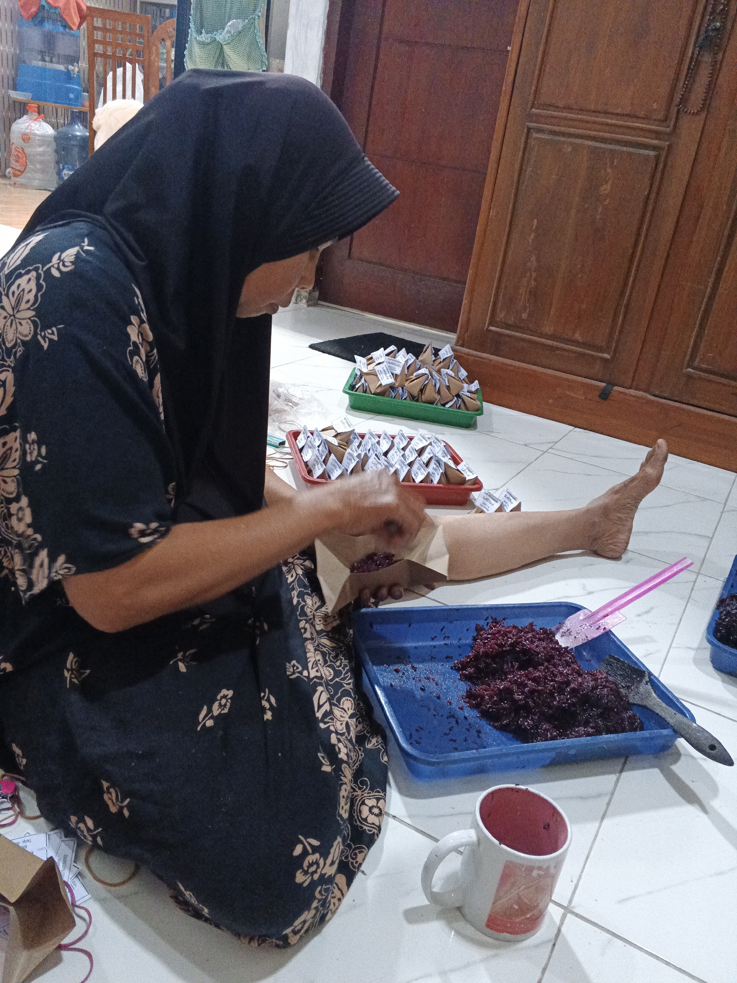 In Kemangsen, Indonesia, a woman wearing a black hijab and a dark patterned dress kneels on a tiled floor, engaged in food preparation. Her hands are occupied with a brown paper bag from which she appears to be pouring a dark red, granular substance. In front of her, a blue plastic tray holds a portion of this same substance, being spread with a small black spatula and a pink plastic tool. Nearby, a white mug with a red interior and patterned exterior sits on the floor. Behind her, small, triangular brown packages with white labels are neatly arranged in red and green plastic containers, suggesting individual servings. The background reveals wooden furniture, including a cabinet with ornate carvings, and water dispenser bottles. The scene is indoors, on a lightly-colored tiled floor, under artificial lighting.