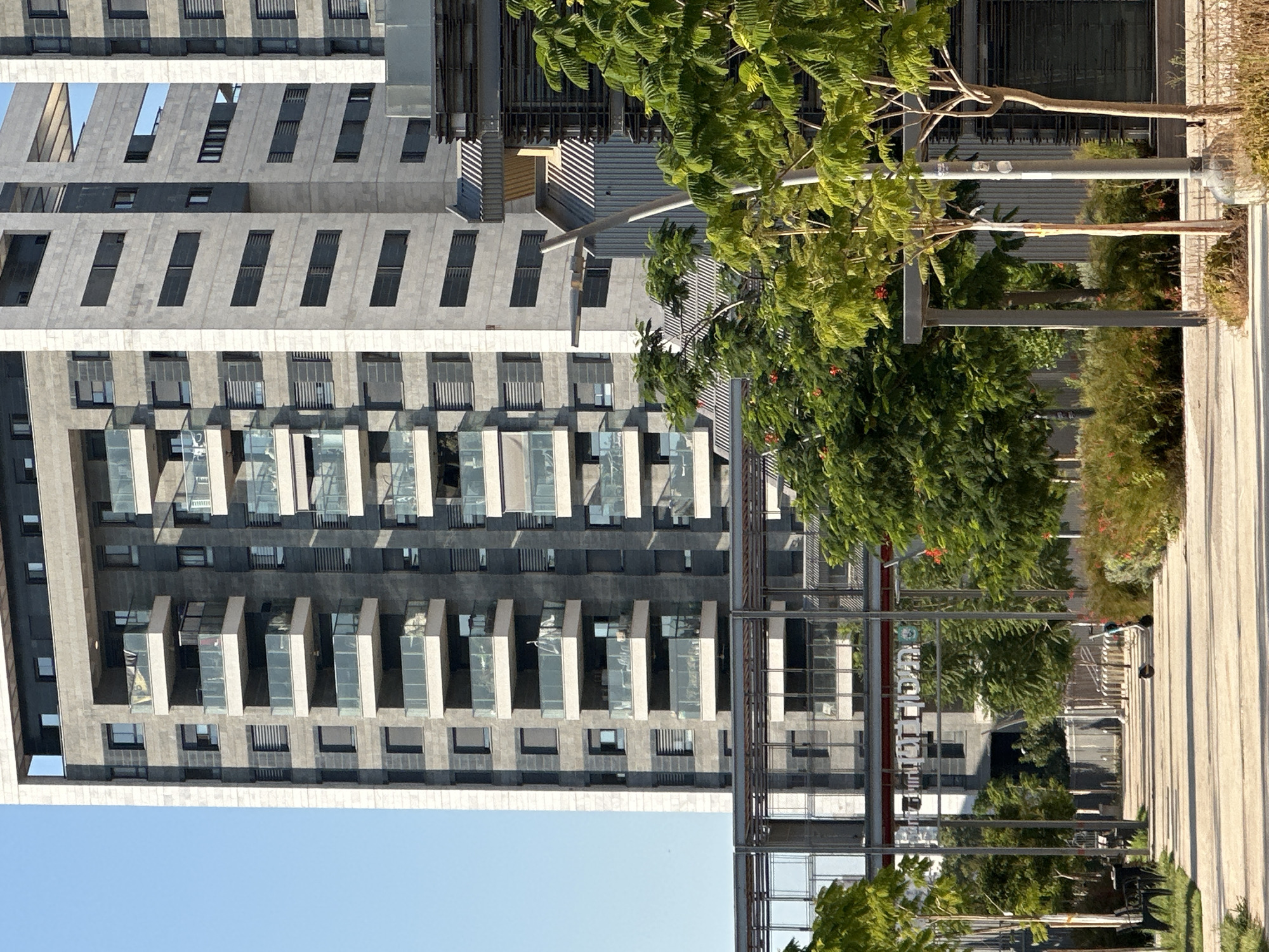 In Ramat Gan, Israel, a modern architectural complex features a tall, grey stone-clad residential building with multiple balconies and dark rectangular windows. Adjacent to it, another building with similar architectural elements is visible. In the foreground, a paved walkway leads through a park with lush green trees and bushes. Several streetlights and a modern sign with Hebrew lettering are present. A bench and a bicycle rack are also visible. The sky is clear and blue.