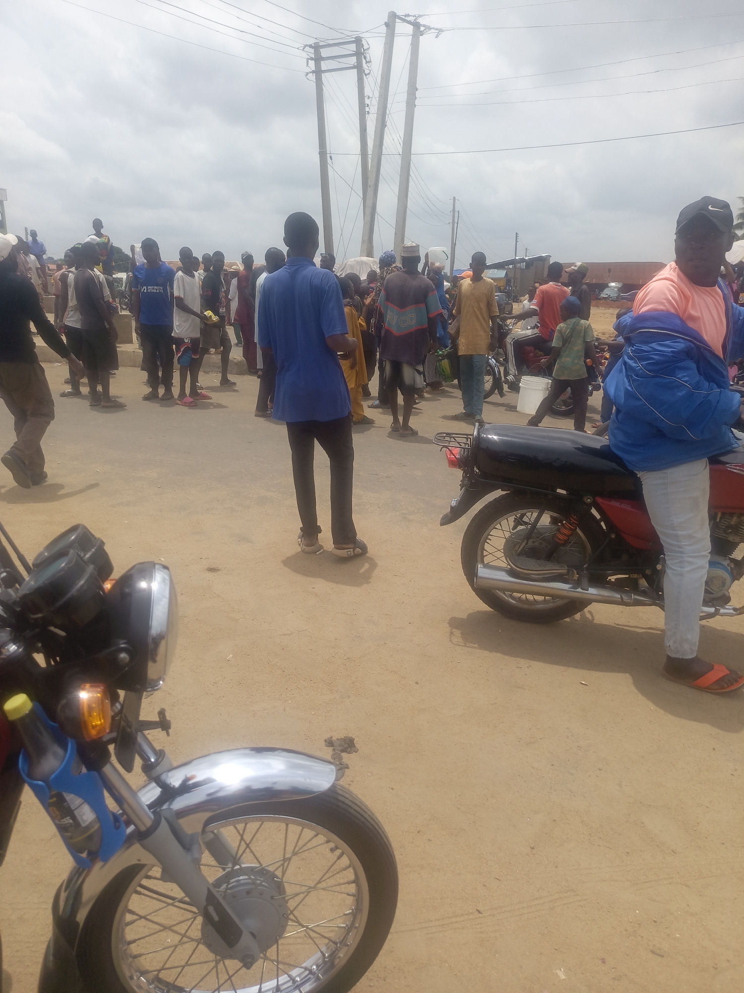 The image captures a scene in Akwanga, Nigeria. A group of people is gathered on a sandy or dusty road under a cloudy sky. Power lines and poles are visible in the background. In the foreground, two motorcycles are parked. The motorcycle on the right has a person sitting on it, wearing light-colored jeans, an orange shirt, and a blue jacket. The person is also wearing orange sandals and a black hat. The other motorcycle, on the left, has a blue bottle holder attached to the front. The people in the background are wearing various clothing. Some are standing and looking in different directions.